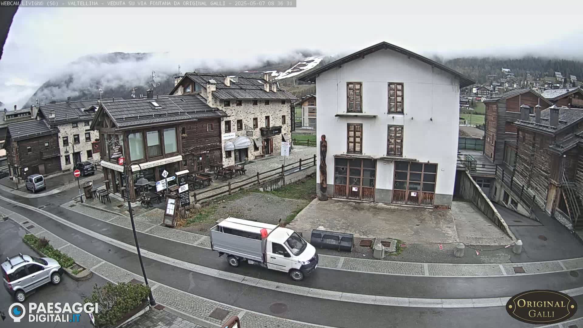 A small village street scene with overcast weather shows a white delivery truck and several parked cars, surrounded by stone buildings and a mountain shrouded in fog in the background.