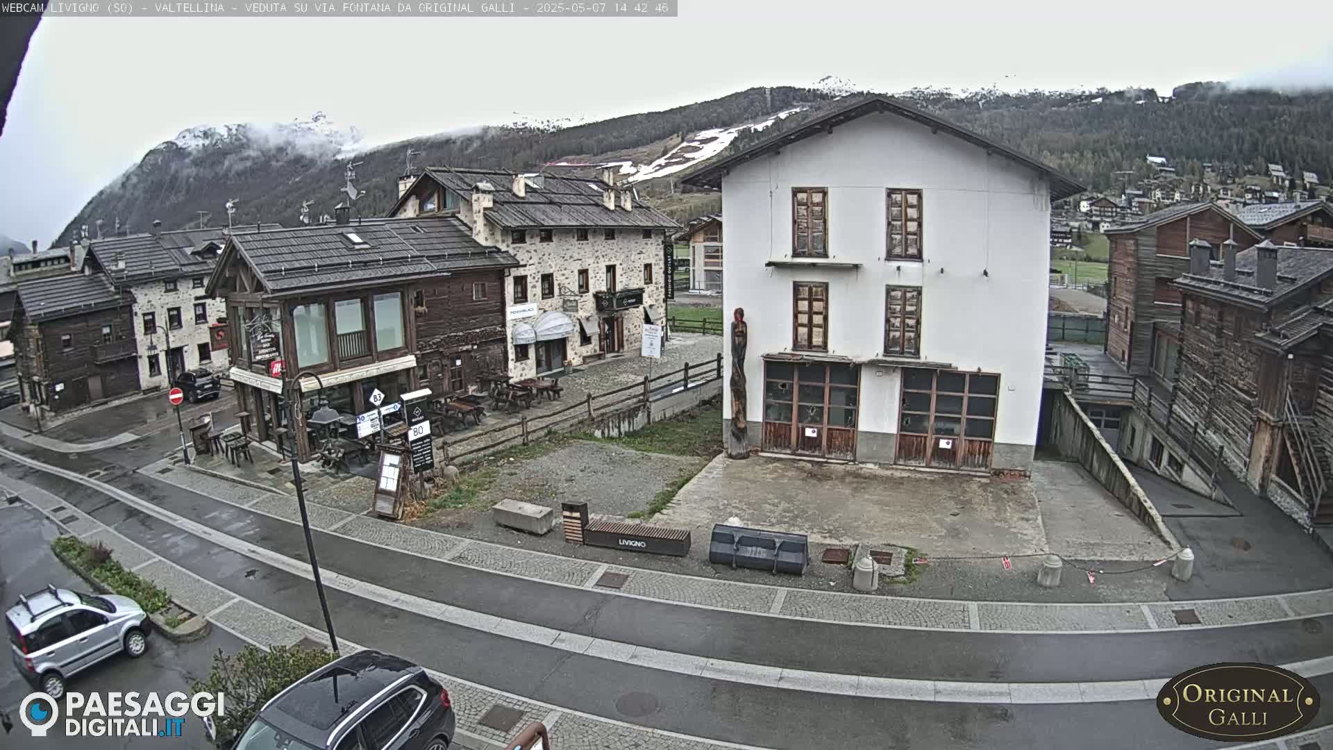 A wet village street scene nestled in a valley, with overcast skies and snow-capped mountains in the background.