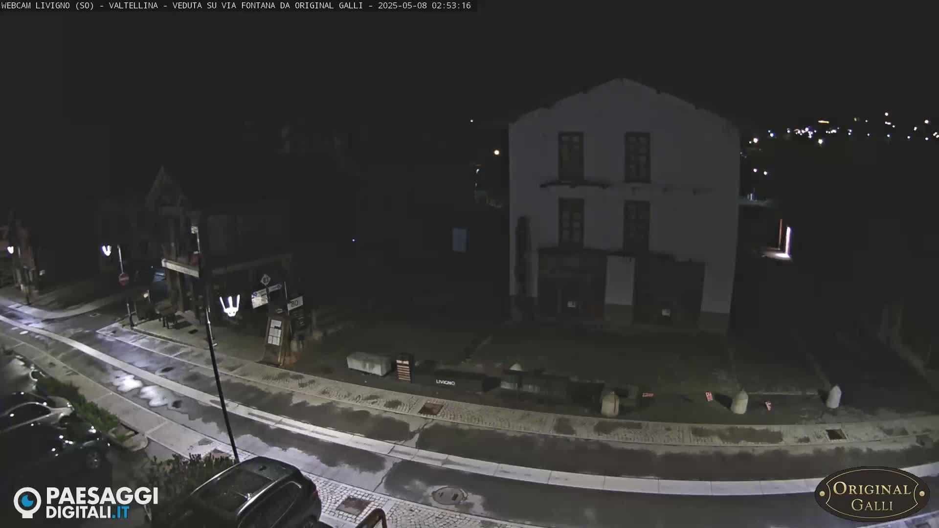 A nighttime view of a wet street curving past buildings in a small town, with a few cars parked along the side.