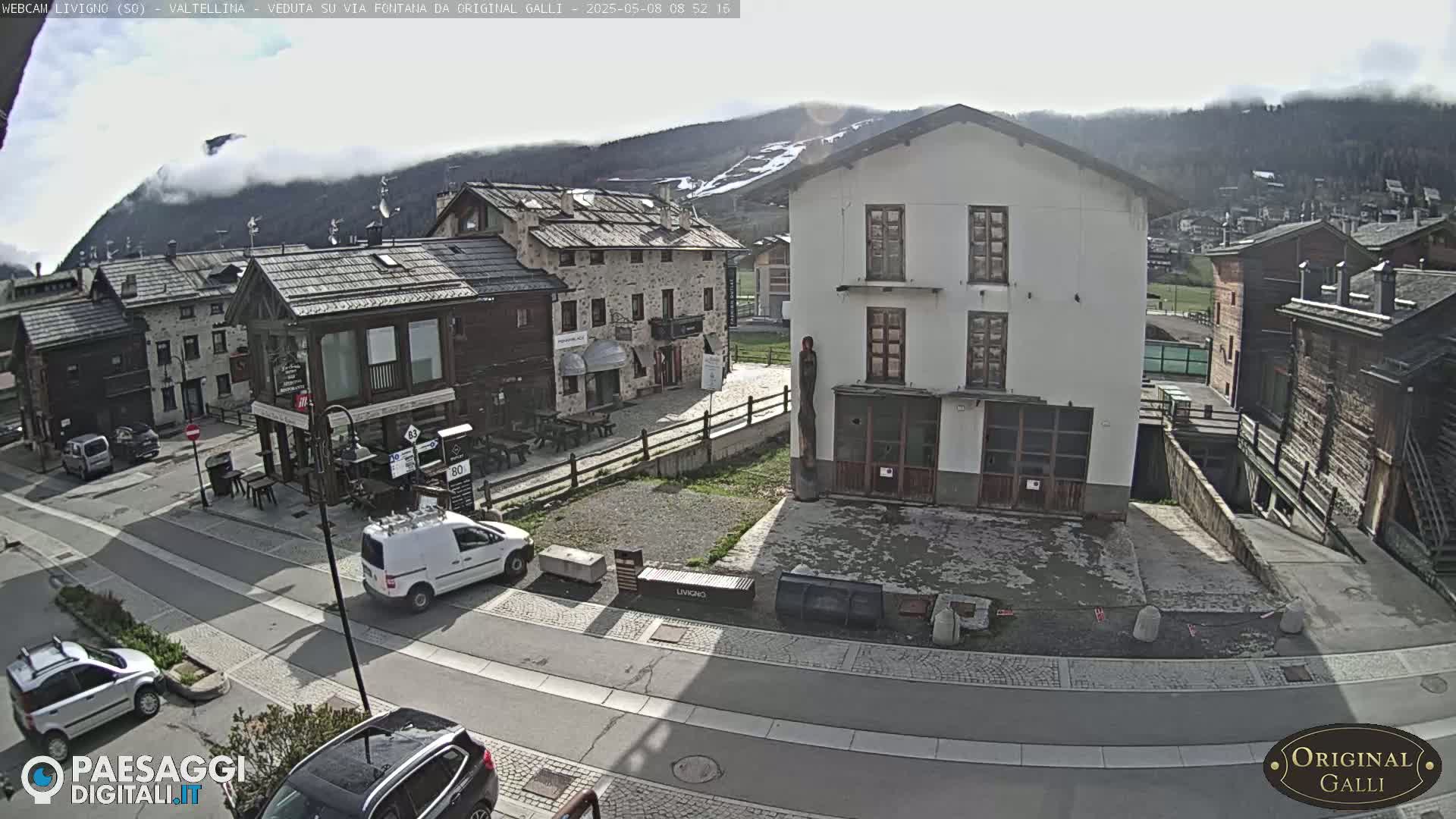 A partly cloudy day reveals a village square with several buildings, a few parked cars, and a van driving away.