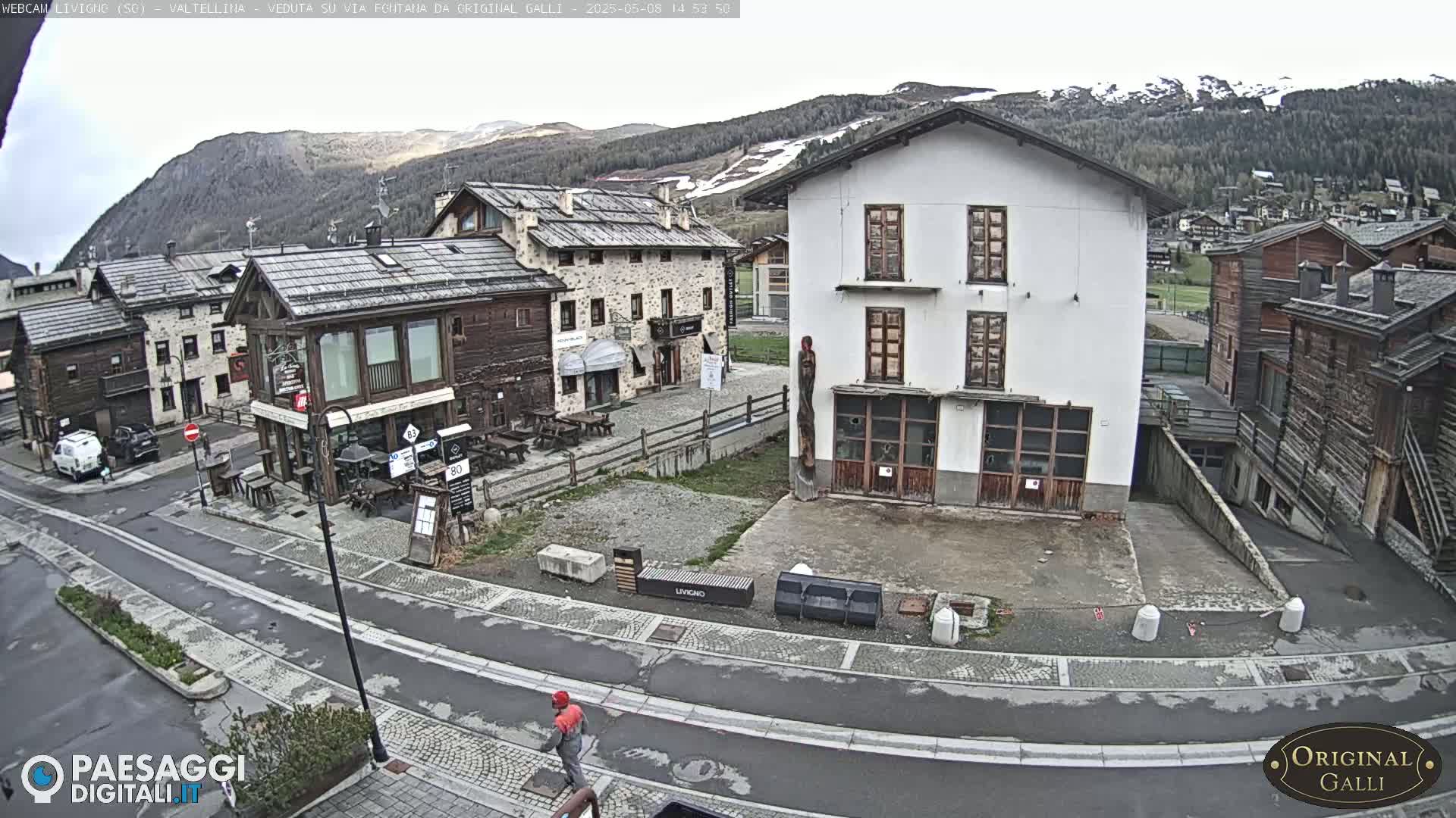 A mostly overcast day reveals a town square with several stone and wood buildings, a few parked cars, and a person walking in the street.