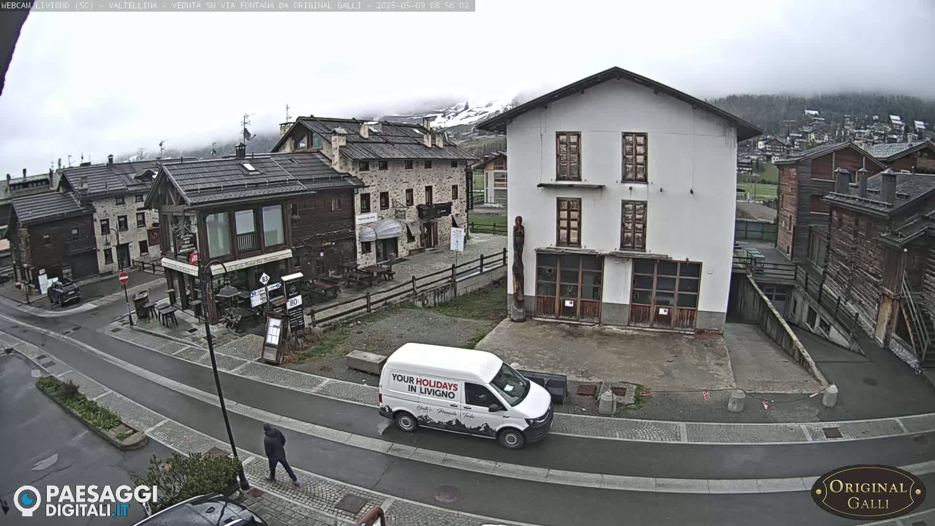 A person walks on a street in a village of stone and wood buildings on an overcast day, with a white van parked nearby.