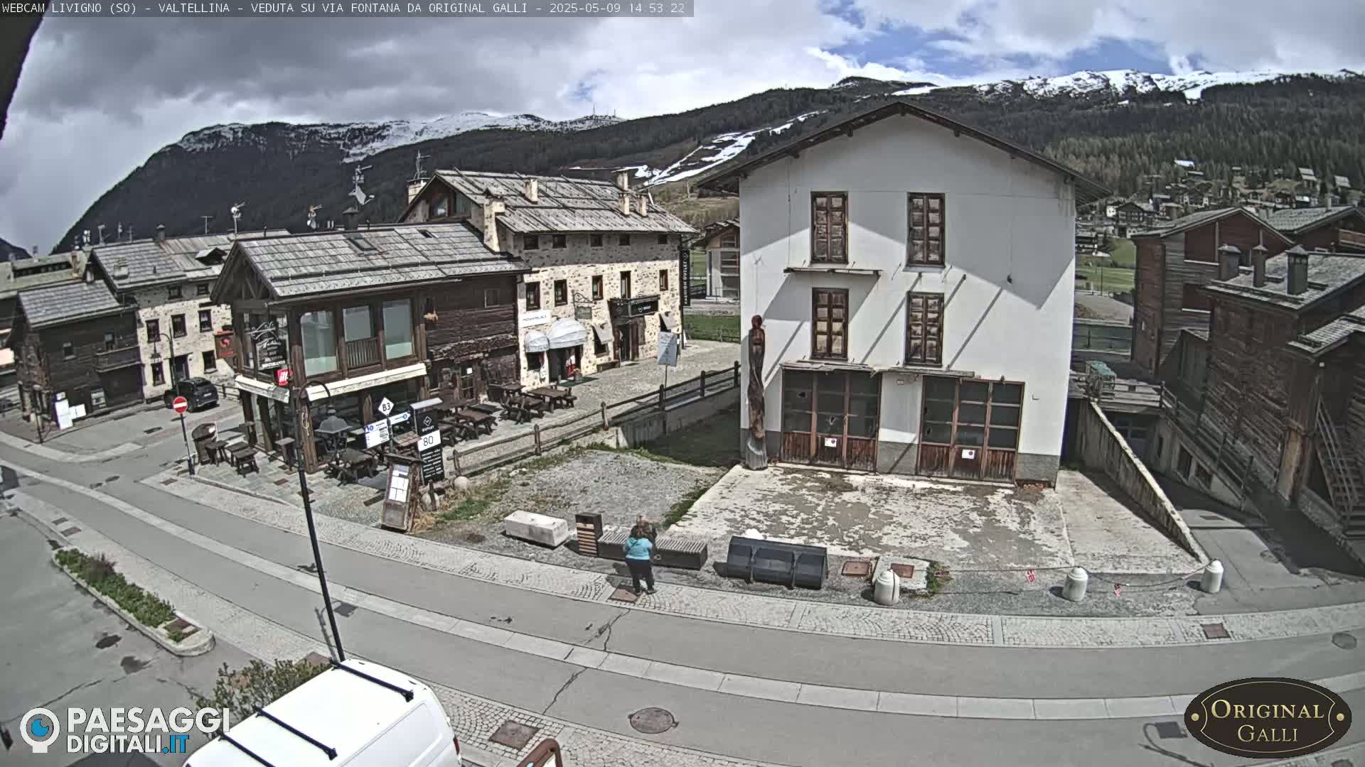 A village square with several buildings, including a white building and several wooden structures, is viewed under partly cloudy skies with mountains in the background, which are partially snow-capped.