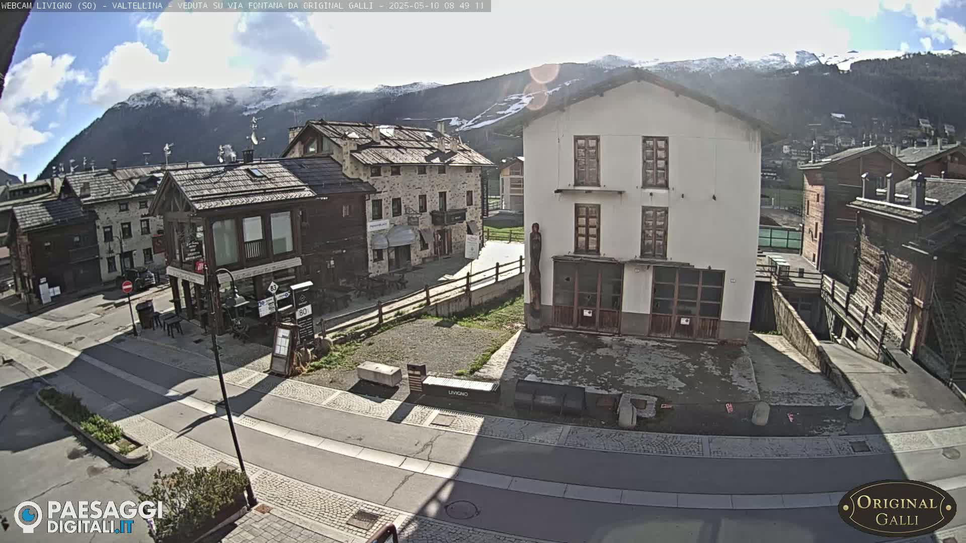 A sunny view of a village square nestled in a valley, with snow-capped mountains in the background.