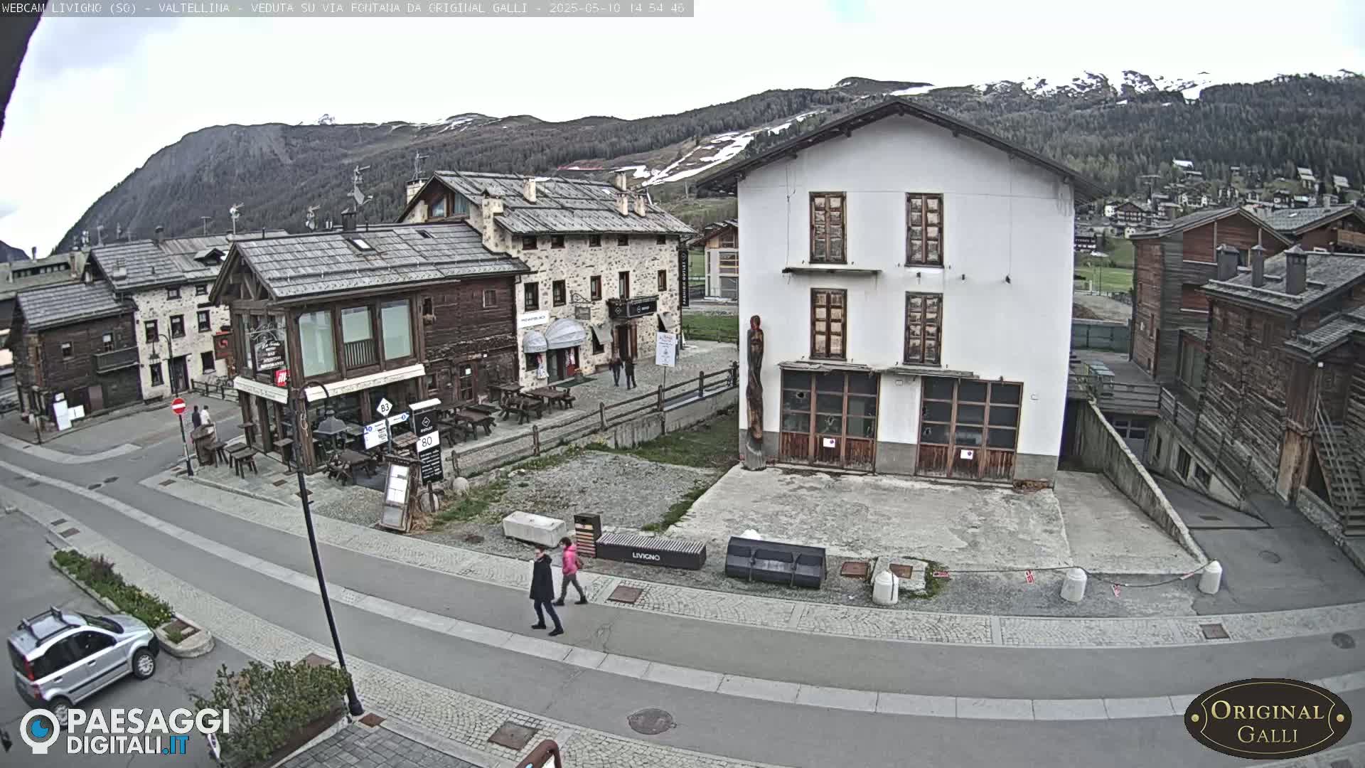 A mostly cloudy day reveals a village street scene nestled amongst mountains, with two pedestrians walking past stone and wood buildings.