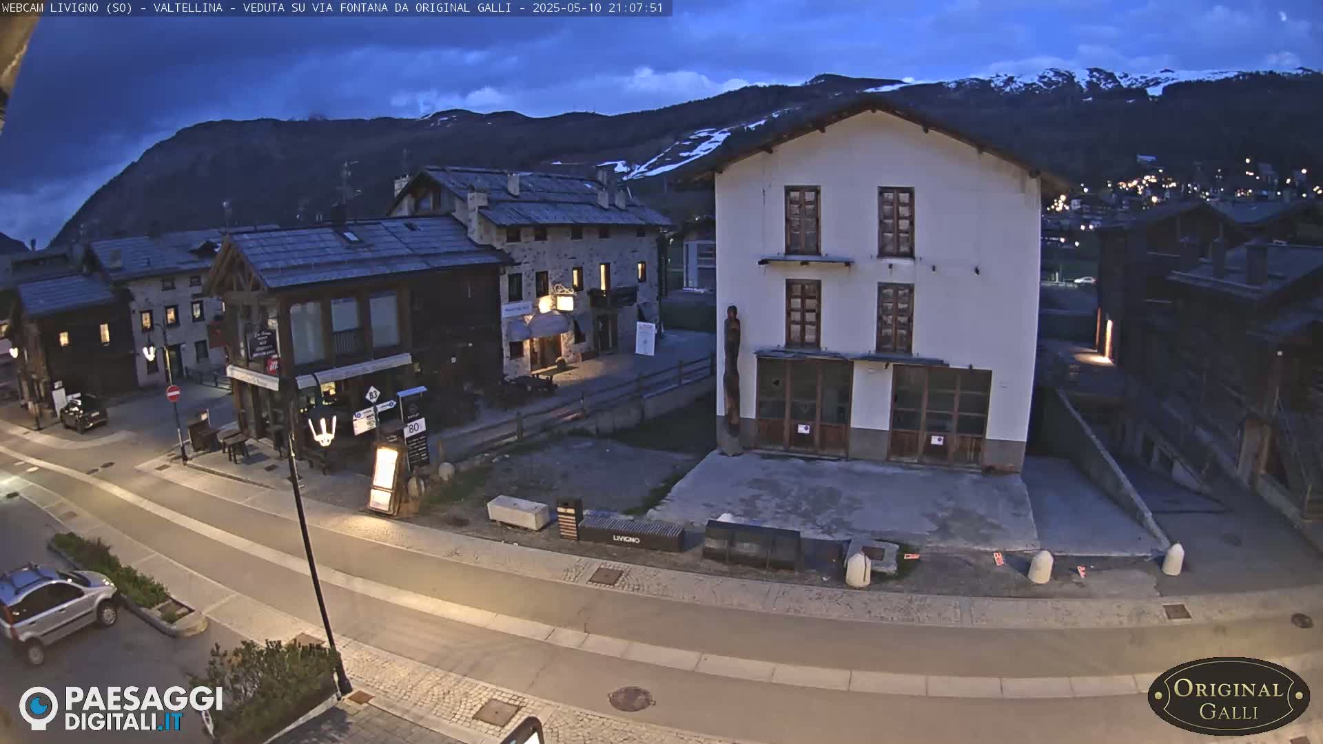 A dimly lit village street scene at twilight, nestled beneath mountains, shows several buildings and a few parked cars.