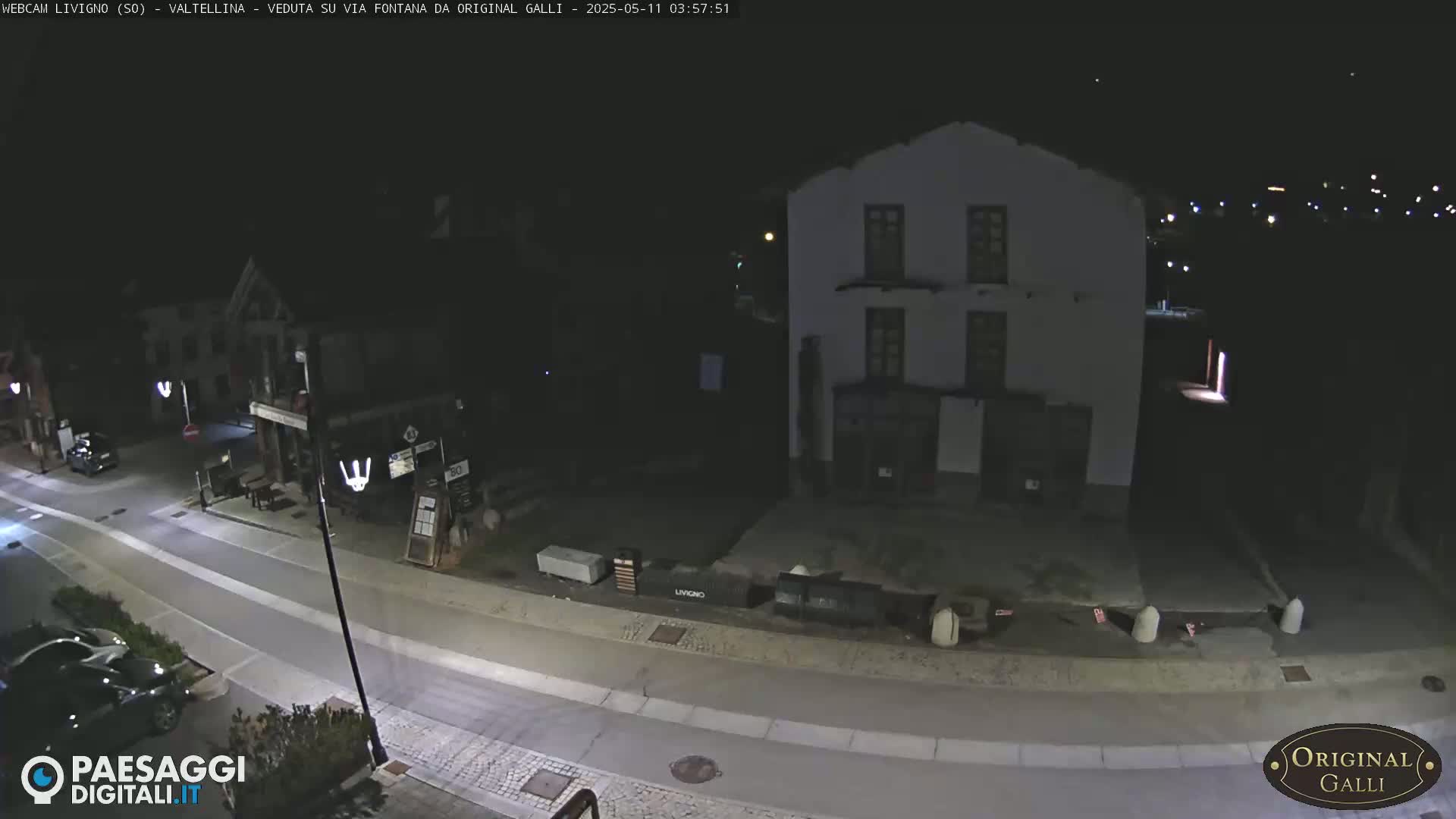 A nighttime, high-angle view of a mostly empty street in a village, with a few parked cars and a large building visible.