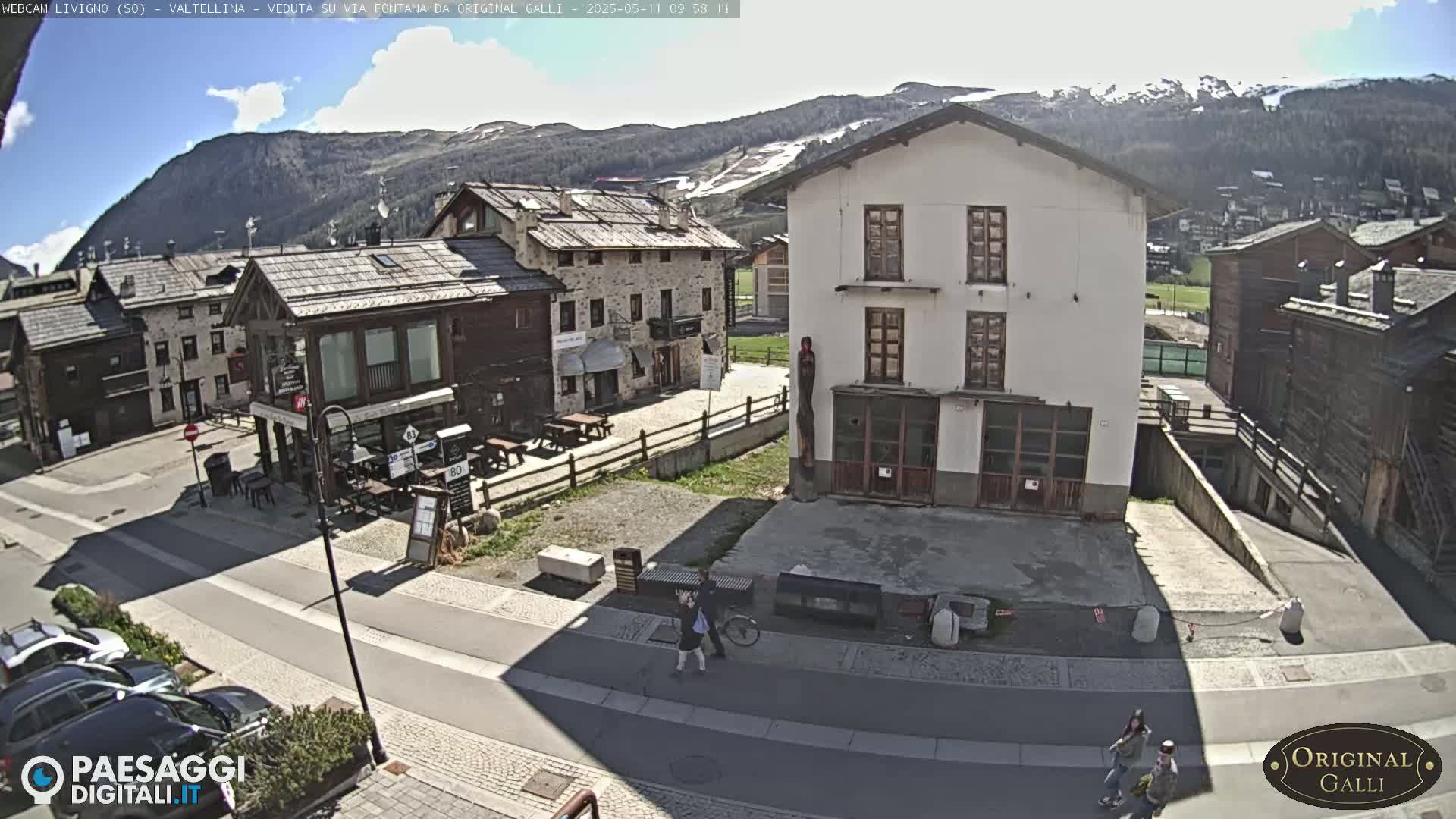 A sunny day in a mountain village shows stone buildings, a paved road, and a few people walking and biking, with mountains in the background.
