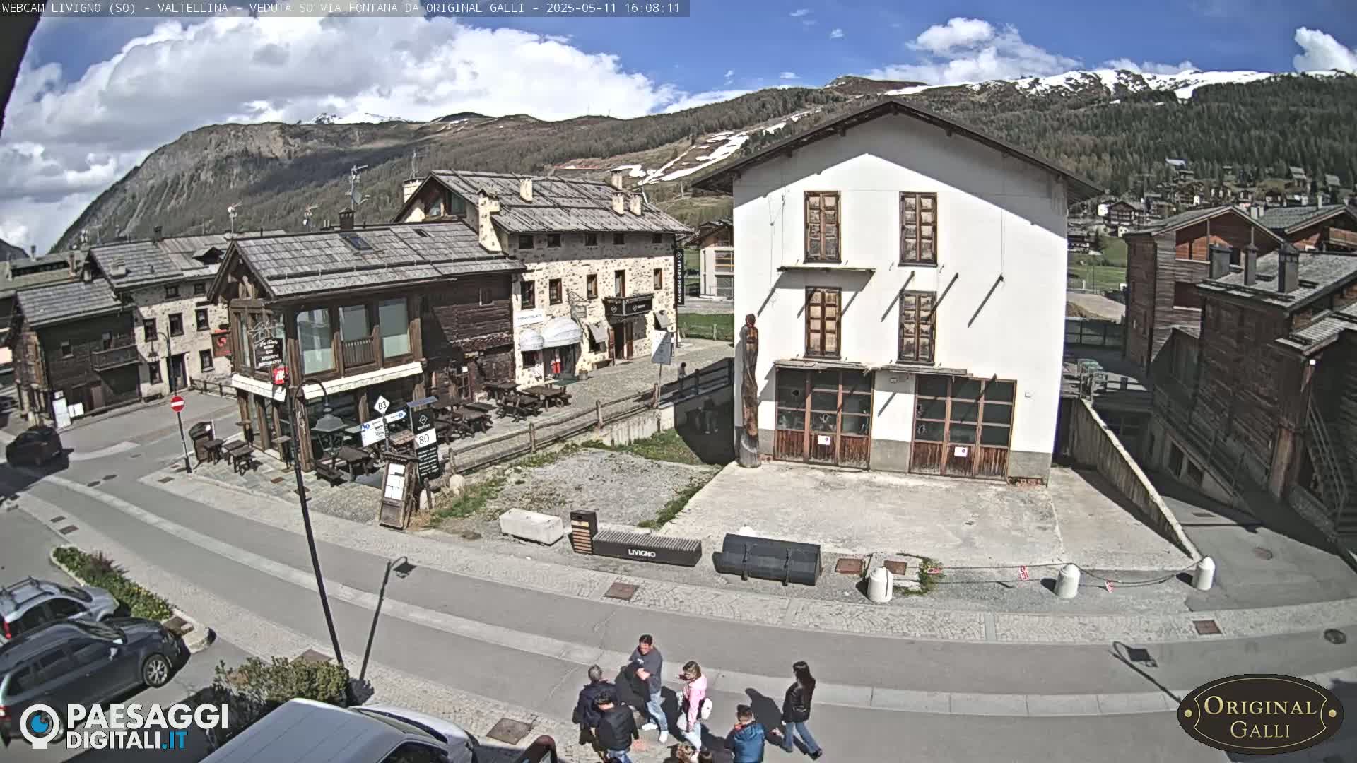 A sunny day in a village nestled in a valley with mountains in the background, showing a street scene with several buildings, parked cars, and a small group of people.