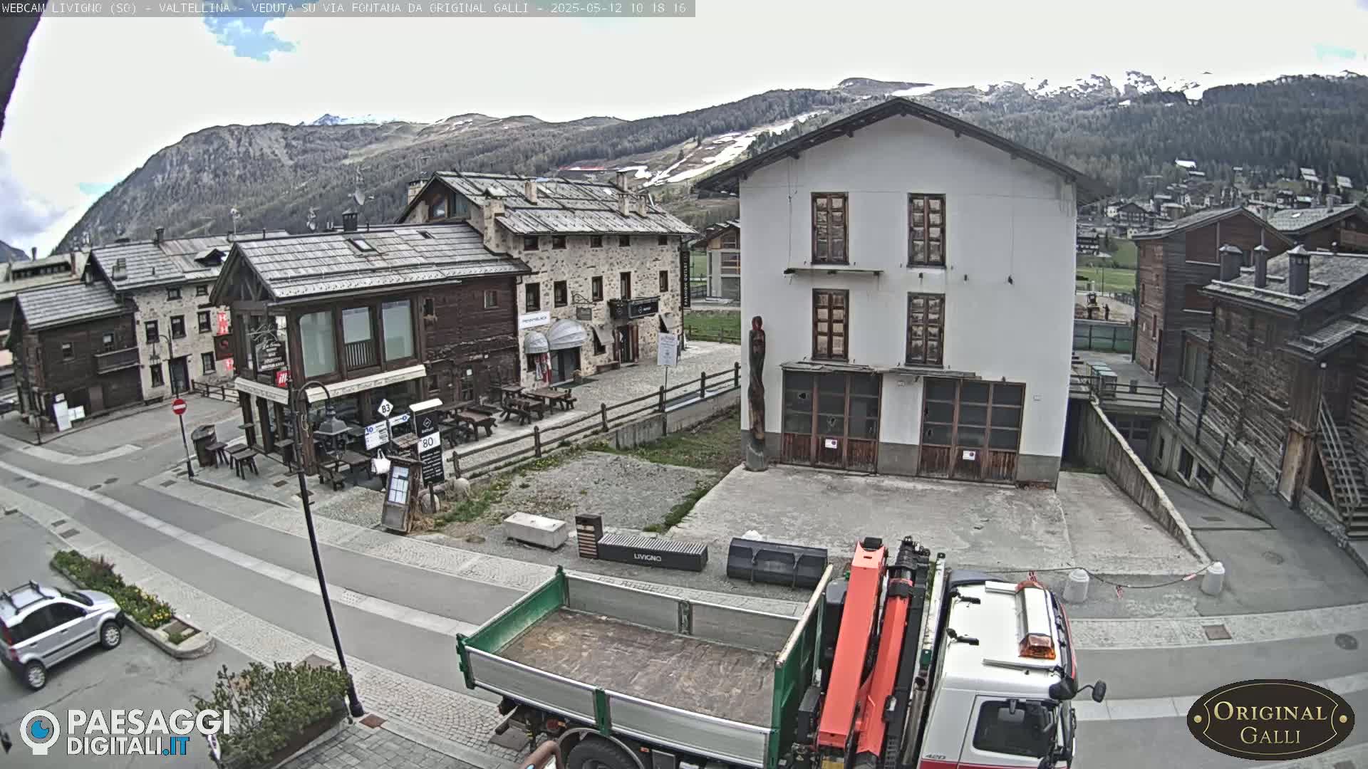 A mostly overcast day reveals a village square in a mountainous region, where a truck with a crane is parked near several stone and wooden buildings.