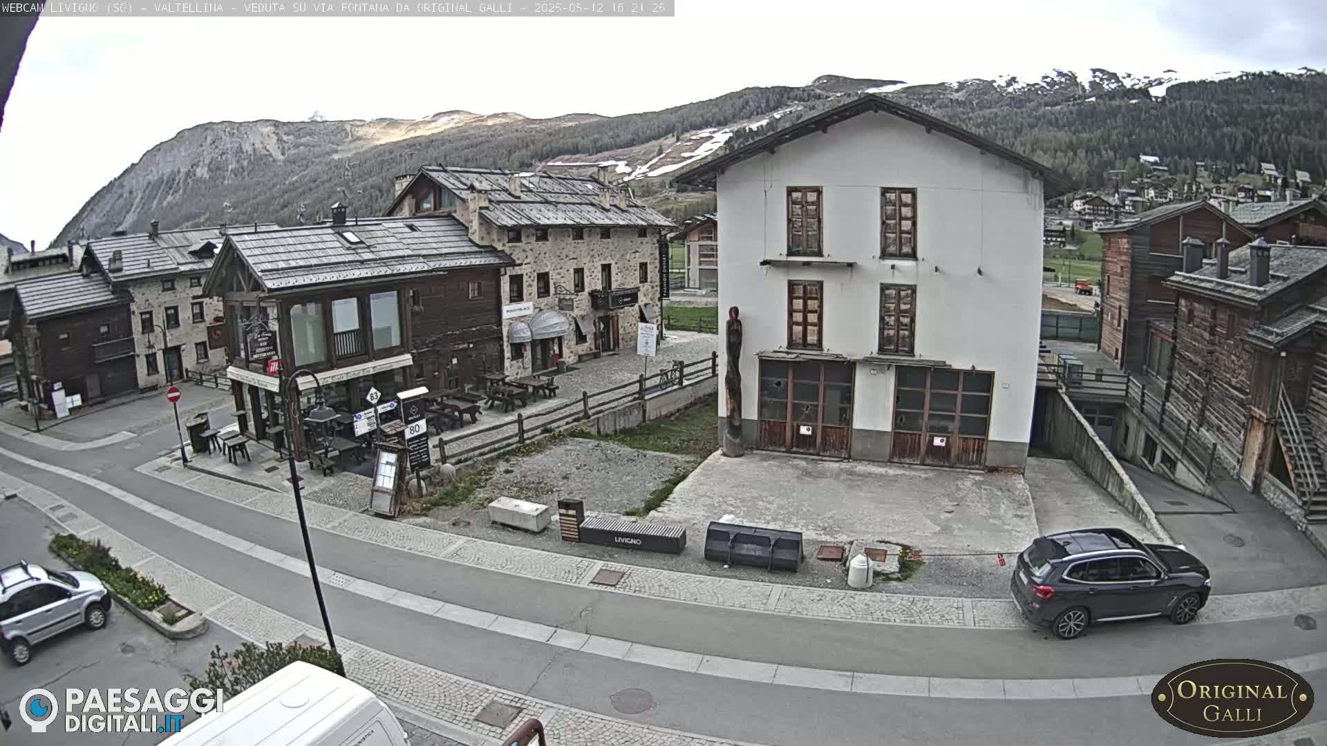 A mostly cloudy day reveals a village nestled in a valley, mountains in the background showing patches of snow.