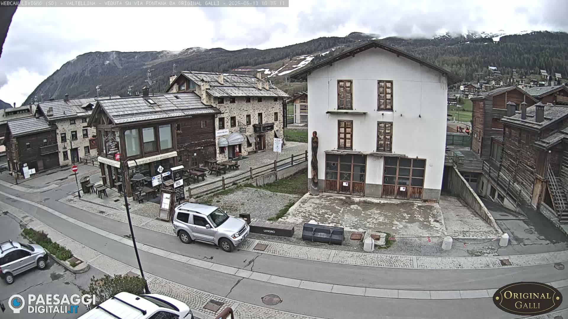A mostly cloudy day reveals a village square in a mountainous region, with several buildings of varying styles and a silver SUV parked near a street corner.