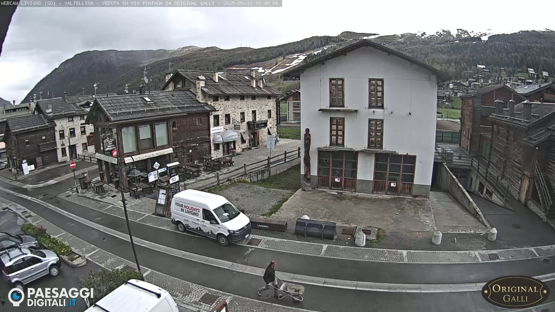 A partly cloudy day reveals a village street scene with several buildings, a white van, and a person wheeling a cart, all set against a backdrop of mountains.