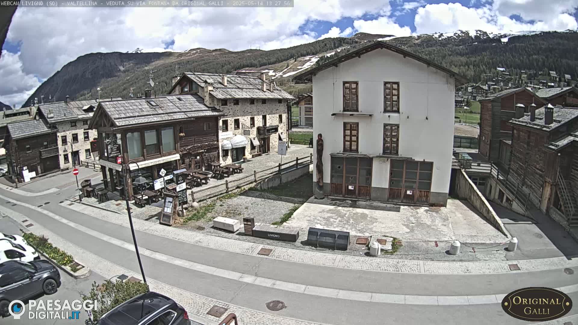A sunny day in a mountain village shows several stone and wood buildings surrounding a paved square with parked cars and a few people.