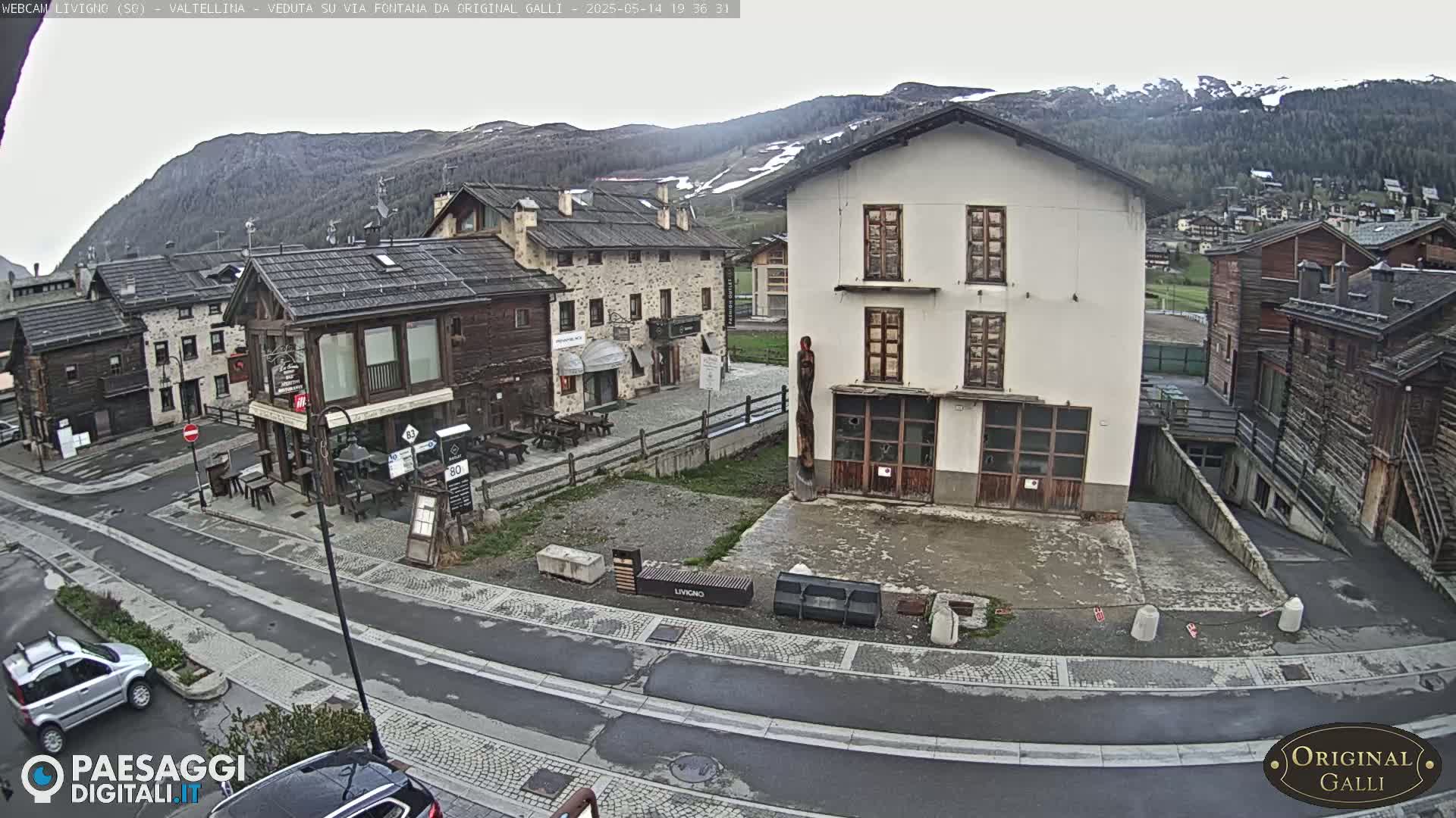A mountain village square with several buildings, a few parked cars, and overcast skies.
