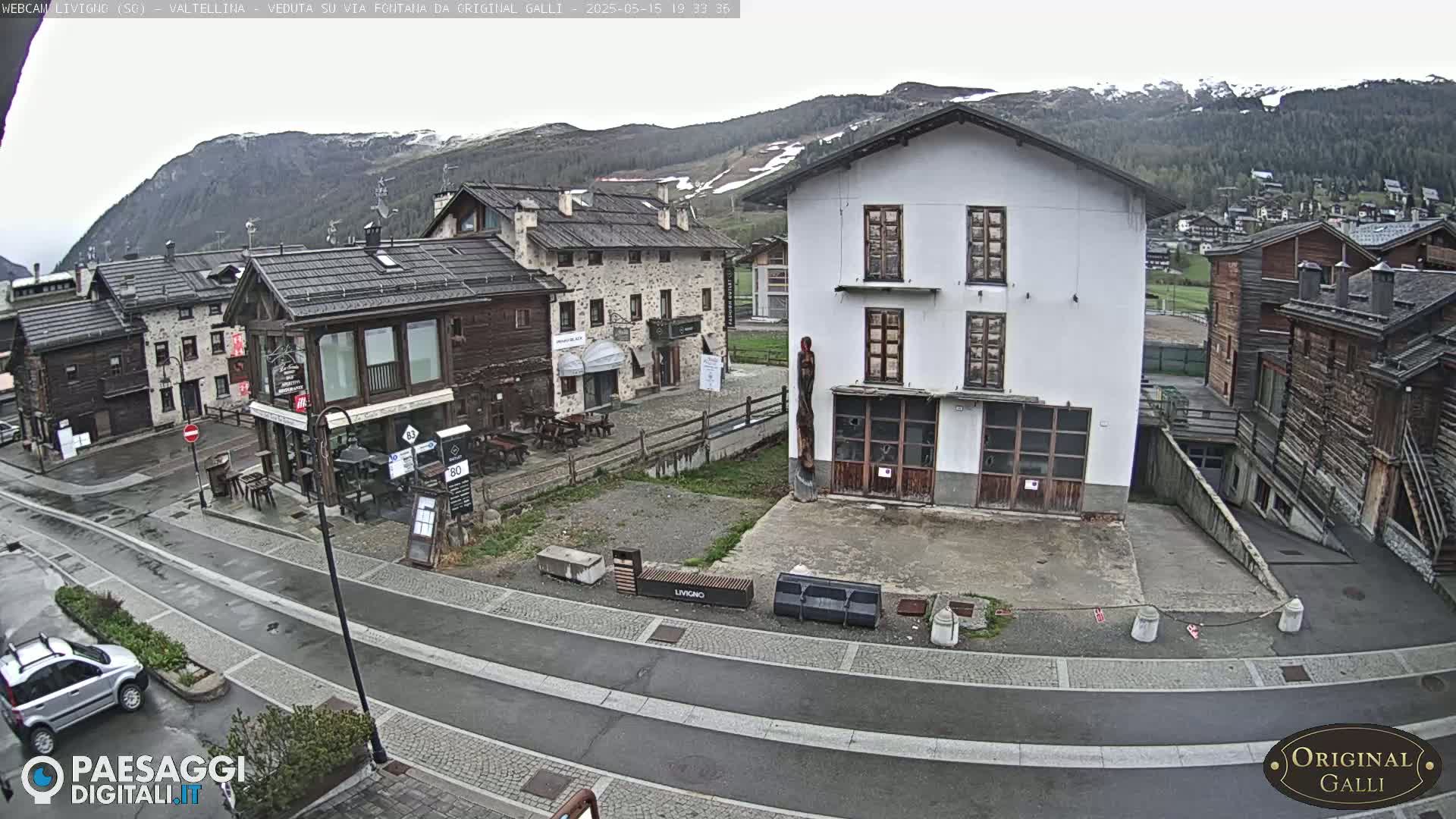 A street scene in a village nestled in a mountain valley shows several buildings, a mostly empty square, and overcast skies.