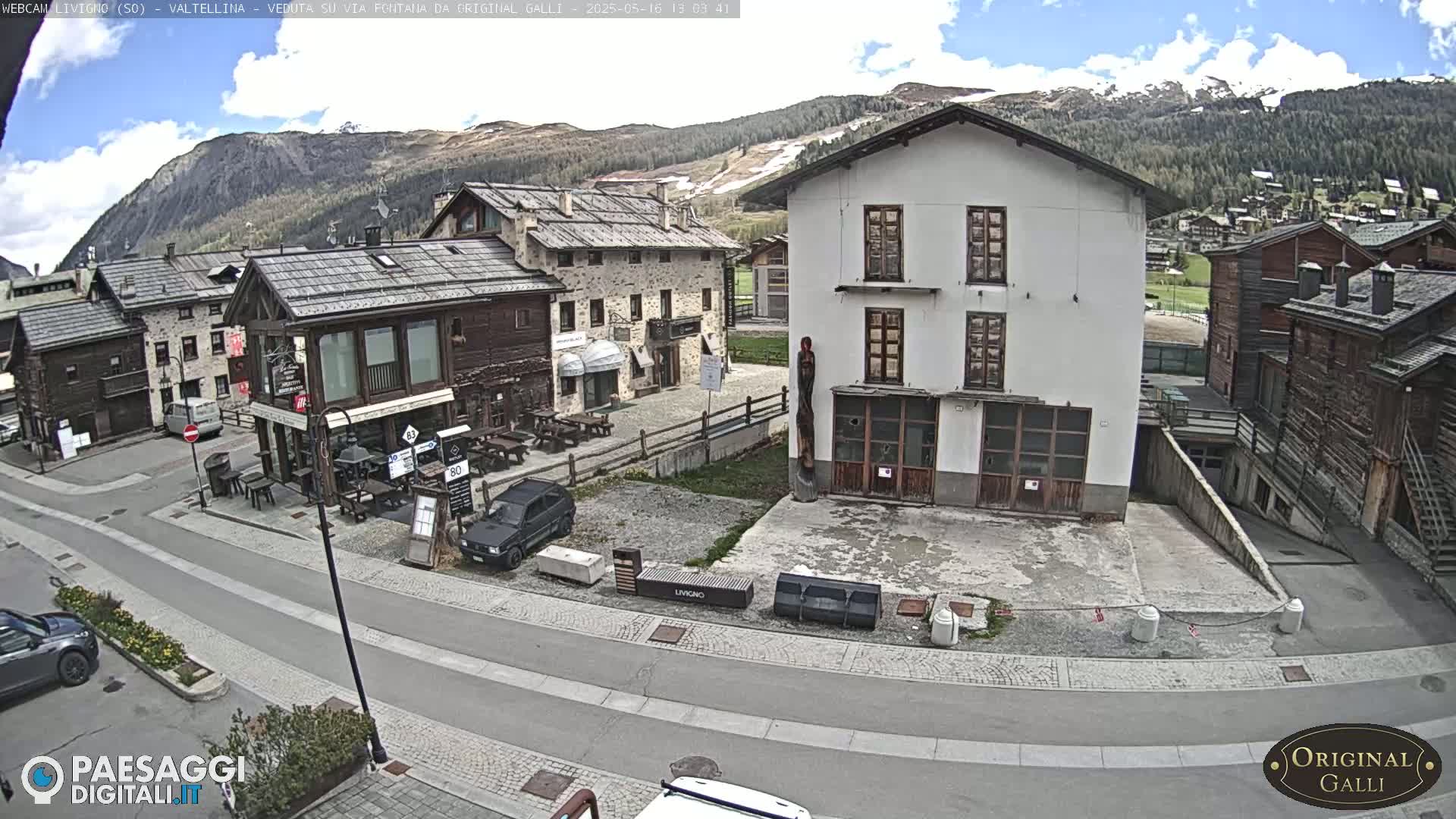 A partly sunny mountain village scene shows several stone and wood buildings surrounding a paved area with parked cars, under a partly cloudy sky.