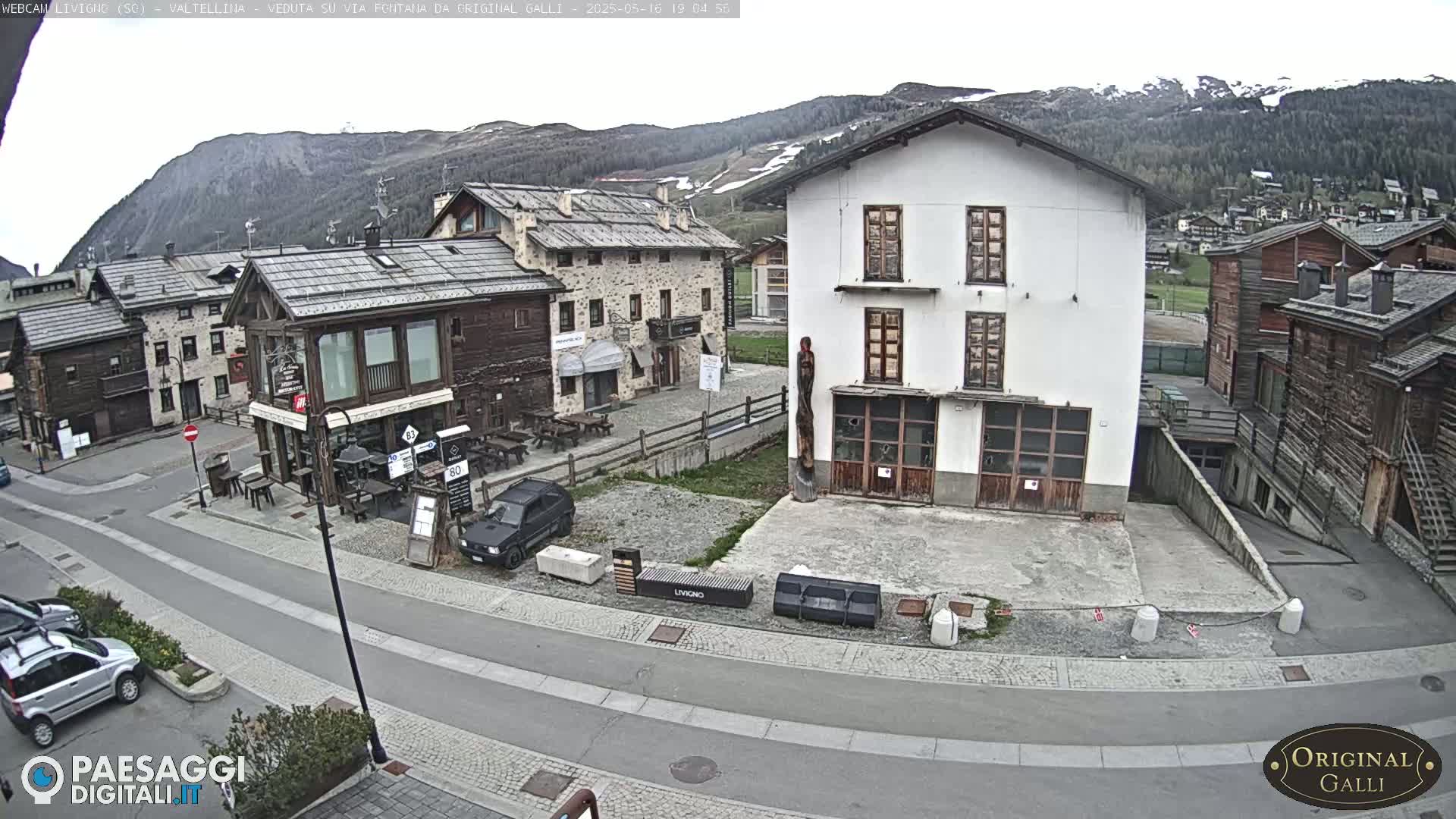 A mostly overcast view of a village nestled in a valley, with mountains in the background and a few cars parked in the street.