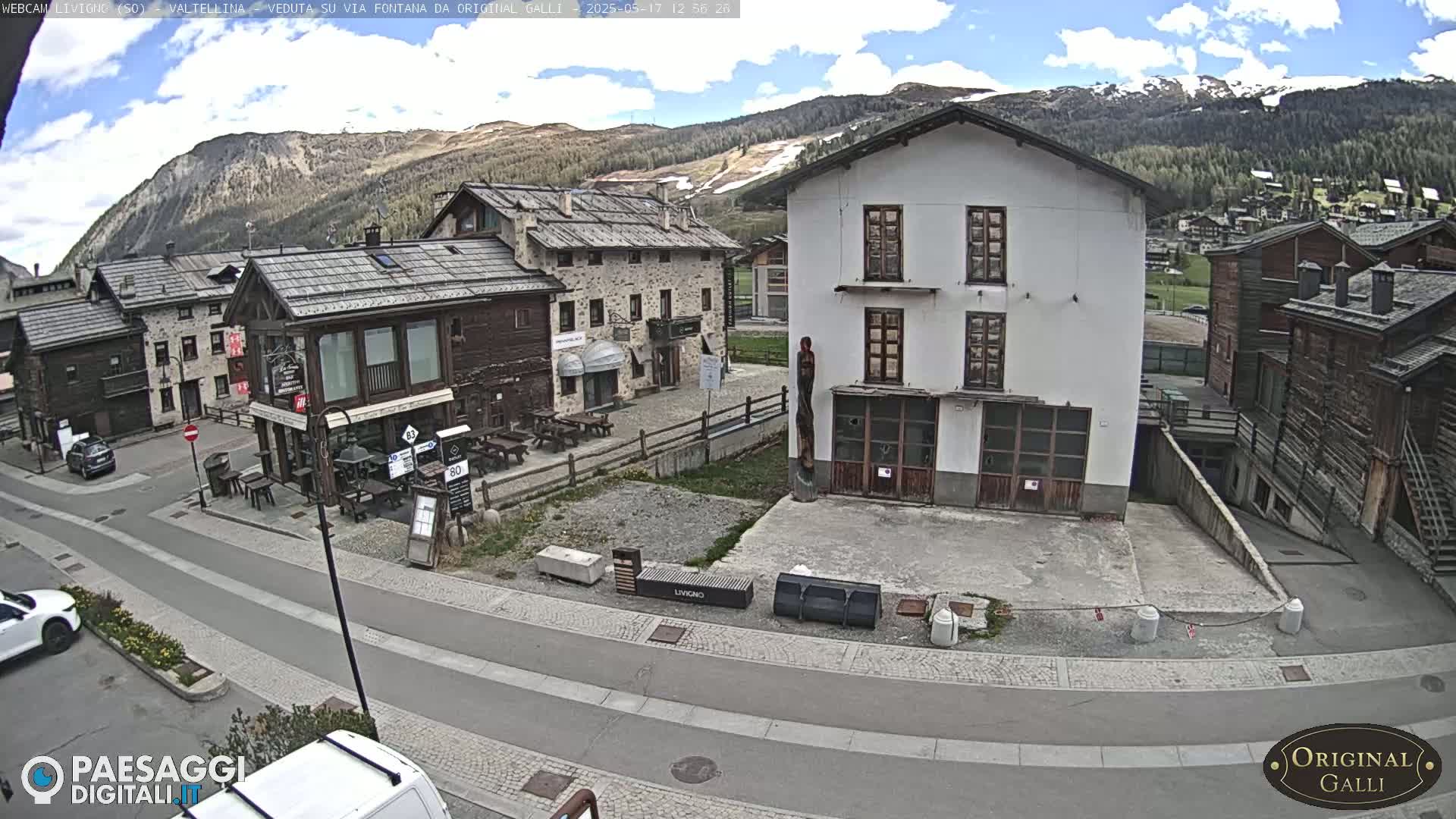 A mostly sunny day reveals a mountain village square with several buildings, including stone and wooden structures, and a few cars parked on a paved road.