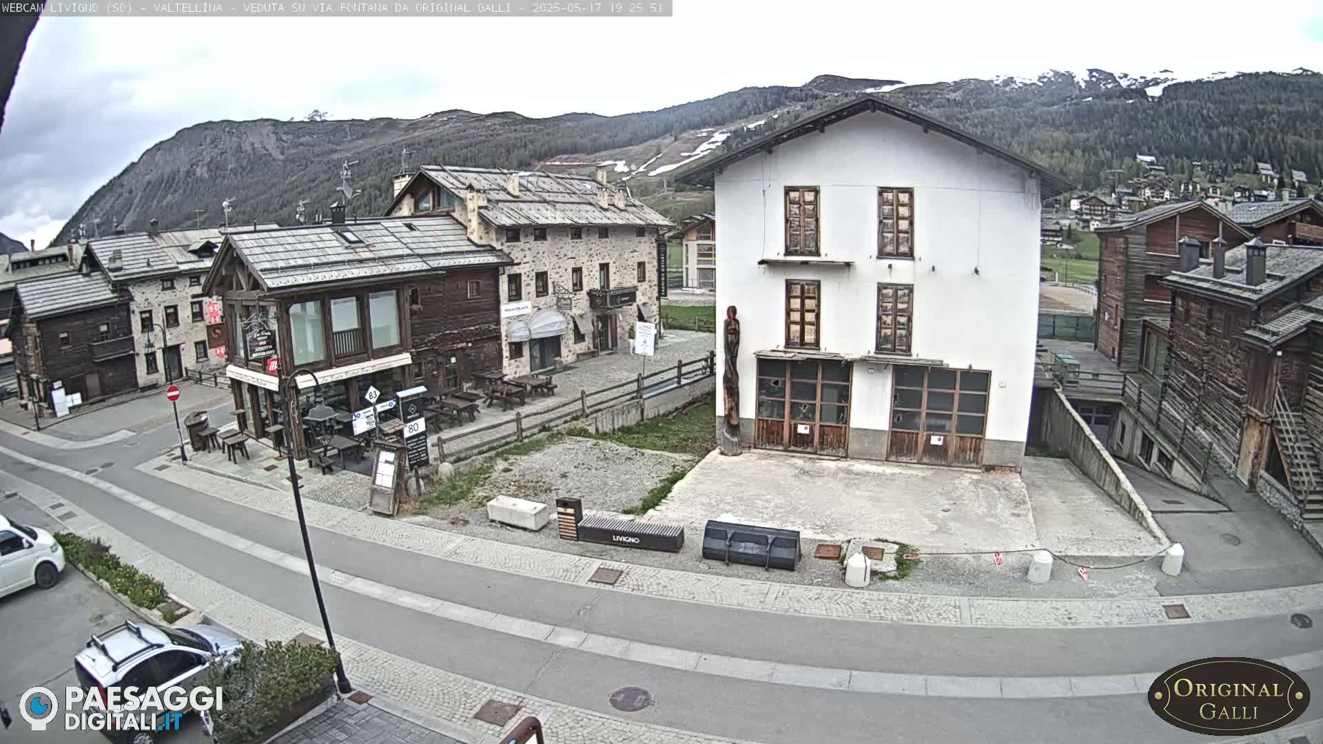 A mountain village street scene under a cloudy sky shows several stone and wood buildings, a paved road, and parked cars.