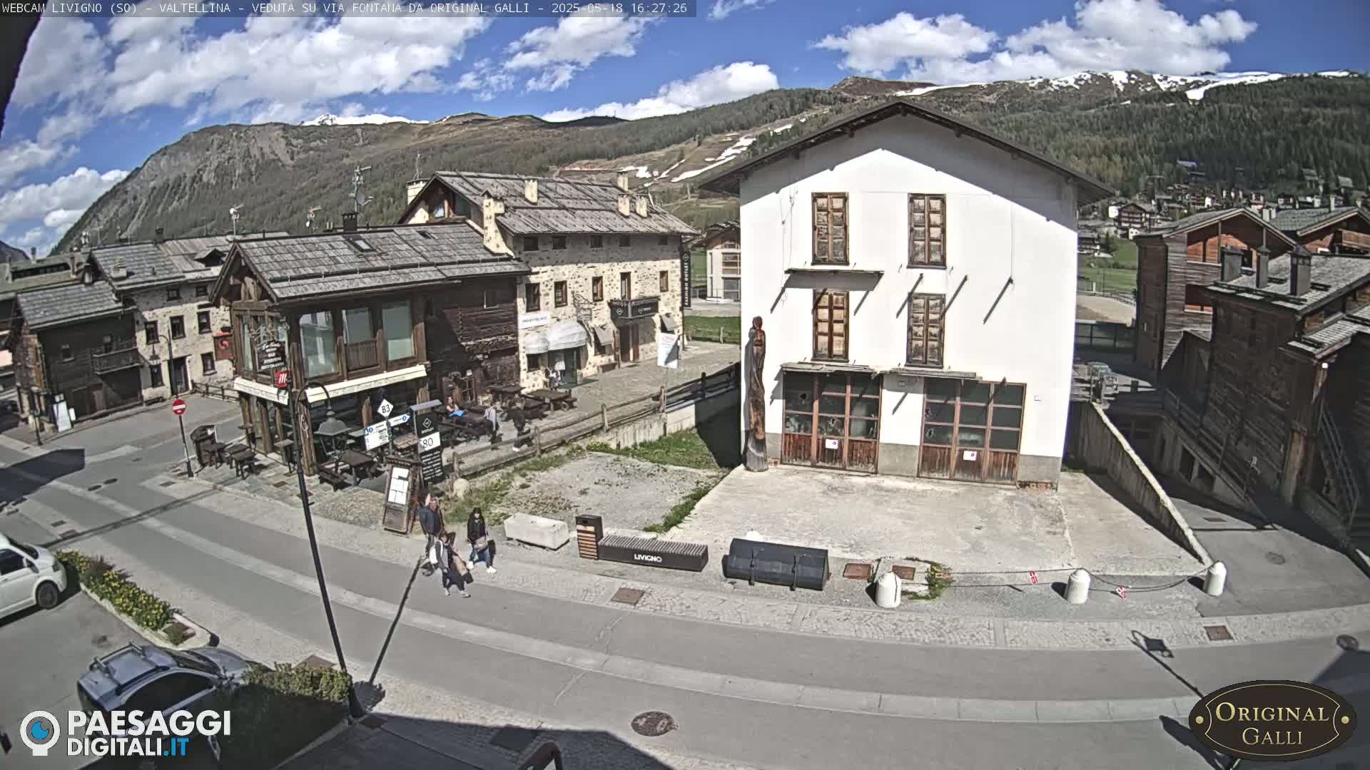 A sunny day in a village nestled in a valley, with mountains in the background and a few people walking around a town square.
