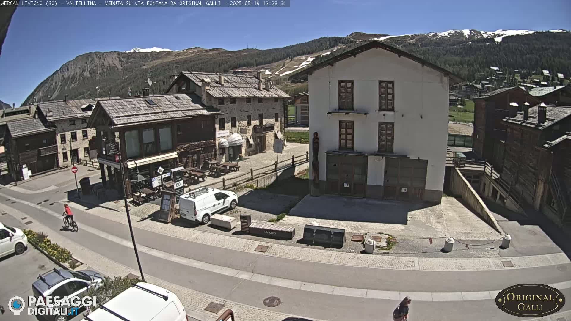 A sunny day reveals a village square with stone buildings, a few parked cars, and a person riding a bicycle, nestled against a backdrop of snow-capped mountains.