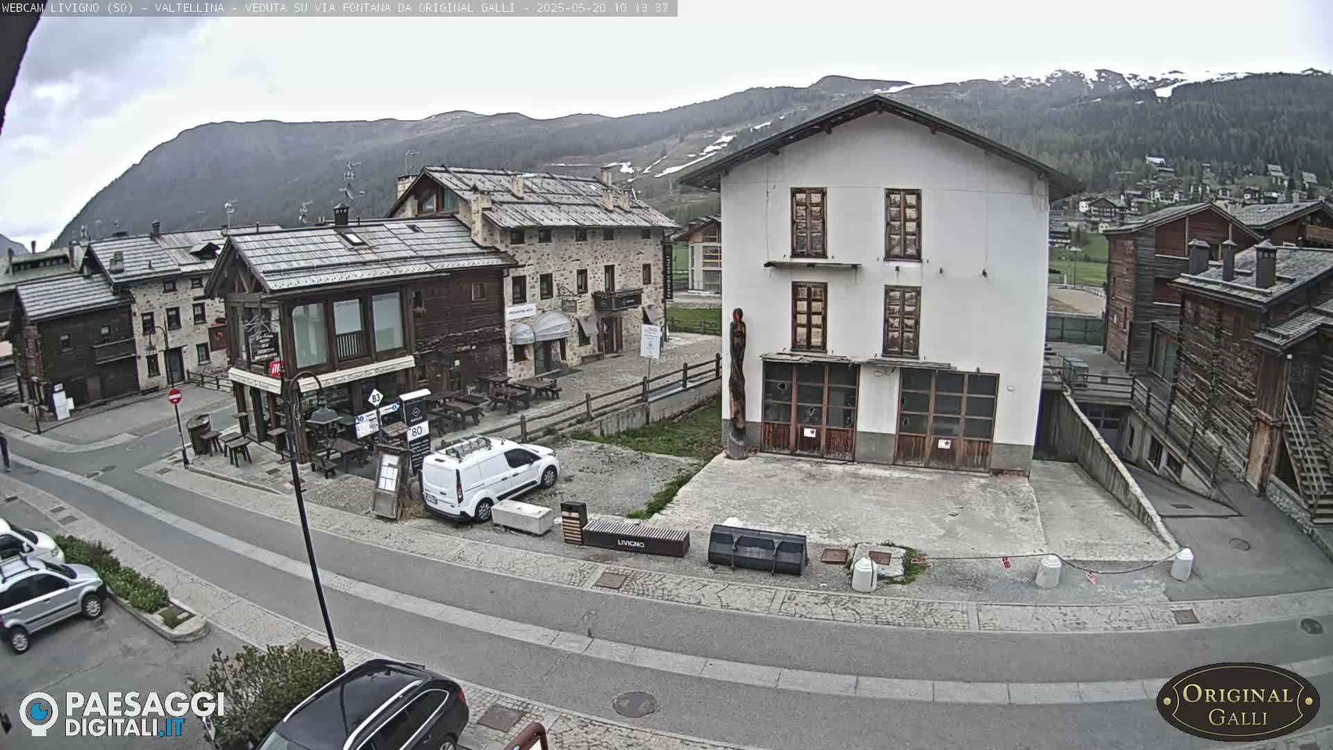 A mostly cloudy day reveals a village scene nestled in a valley, with several buildings of varying architectural styles, a few cars parked, and mountains in the background.