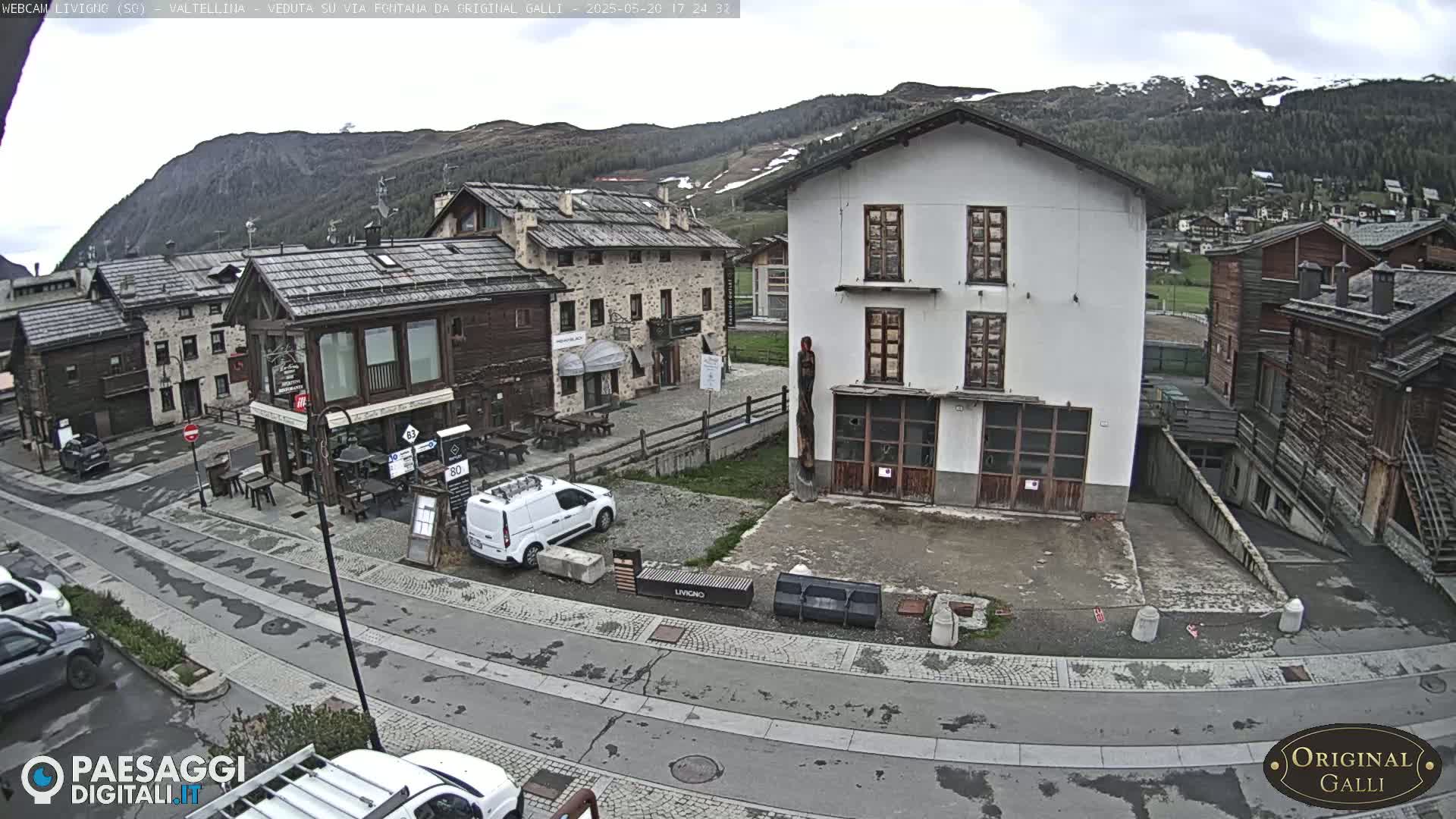 A mostly cloudy day reveals a village scene nestled in a valley, with several buildings of varying architectural styles and a few parked cars visible.