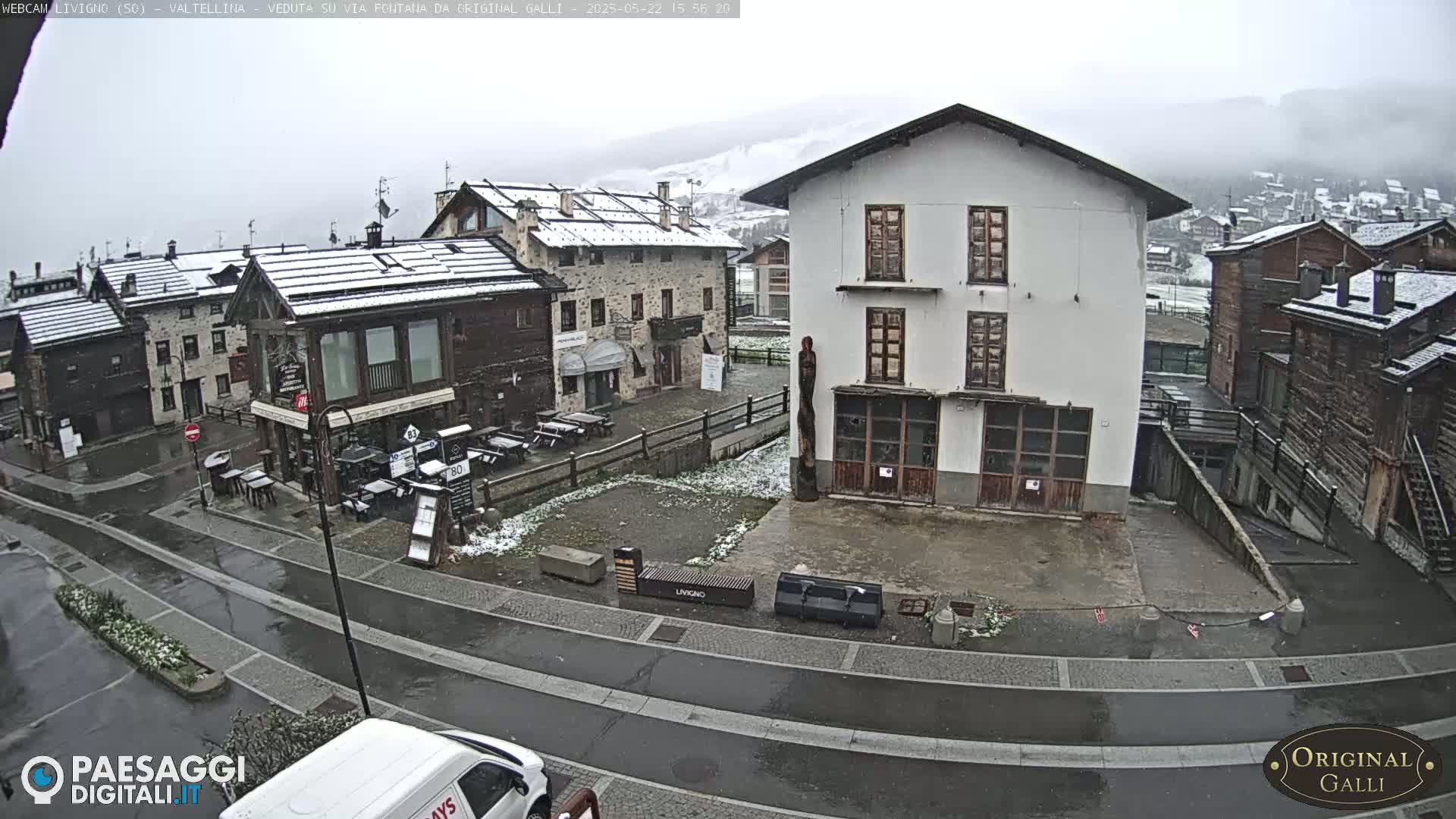 A snow-dusted village square is seen, with several buildings, outdoor seating areas, and a street, under a light snowfall.