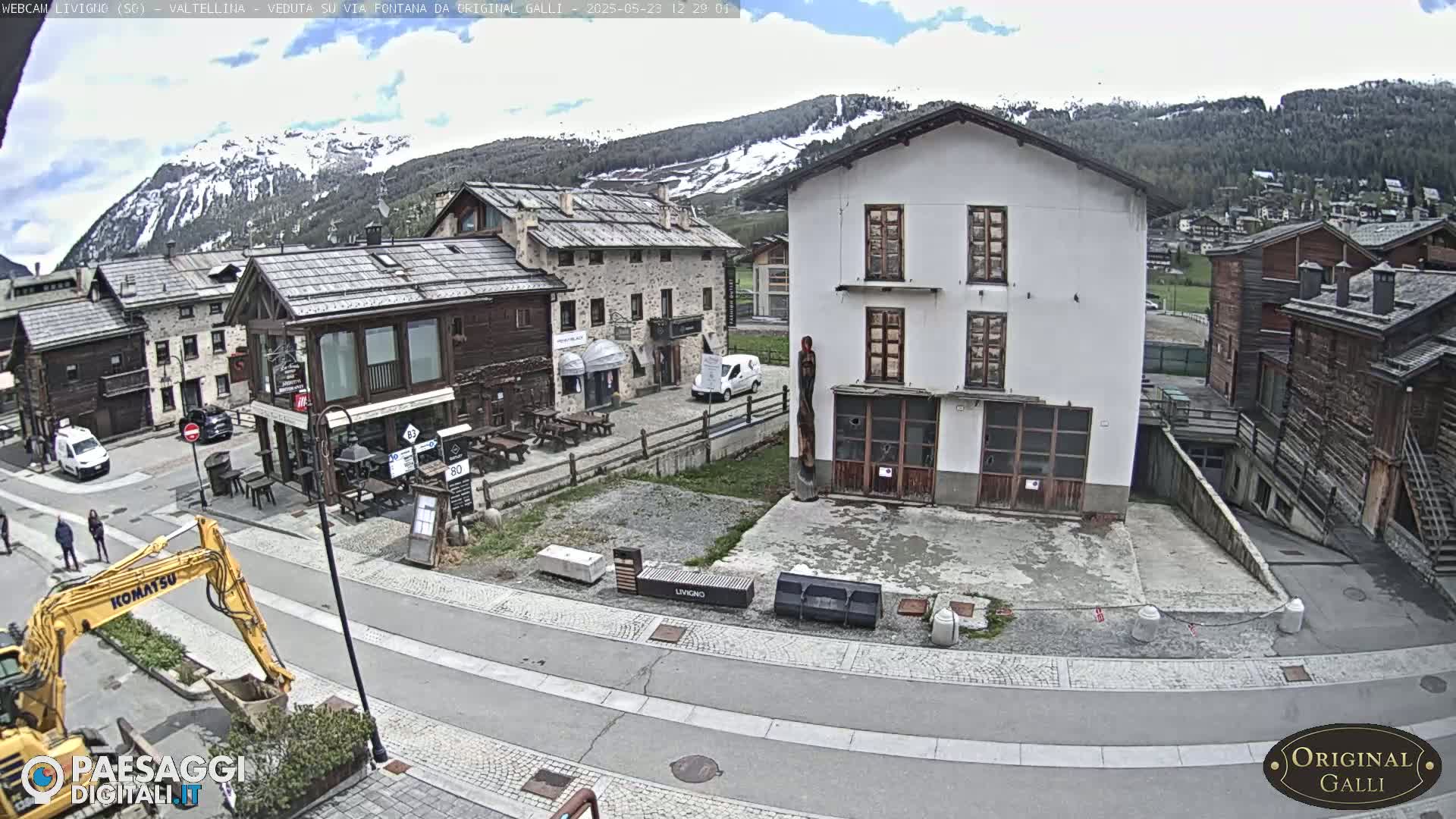 A partly cloudy day in a mountain village with several buildings, a small road, a construction vehicle, and a few people.
