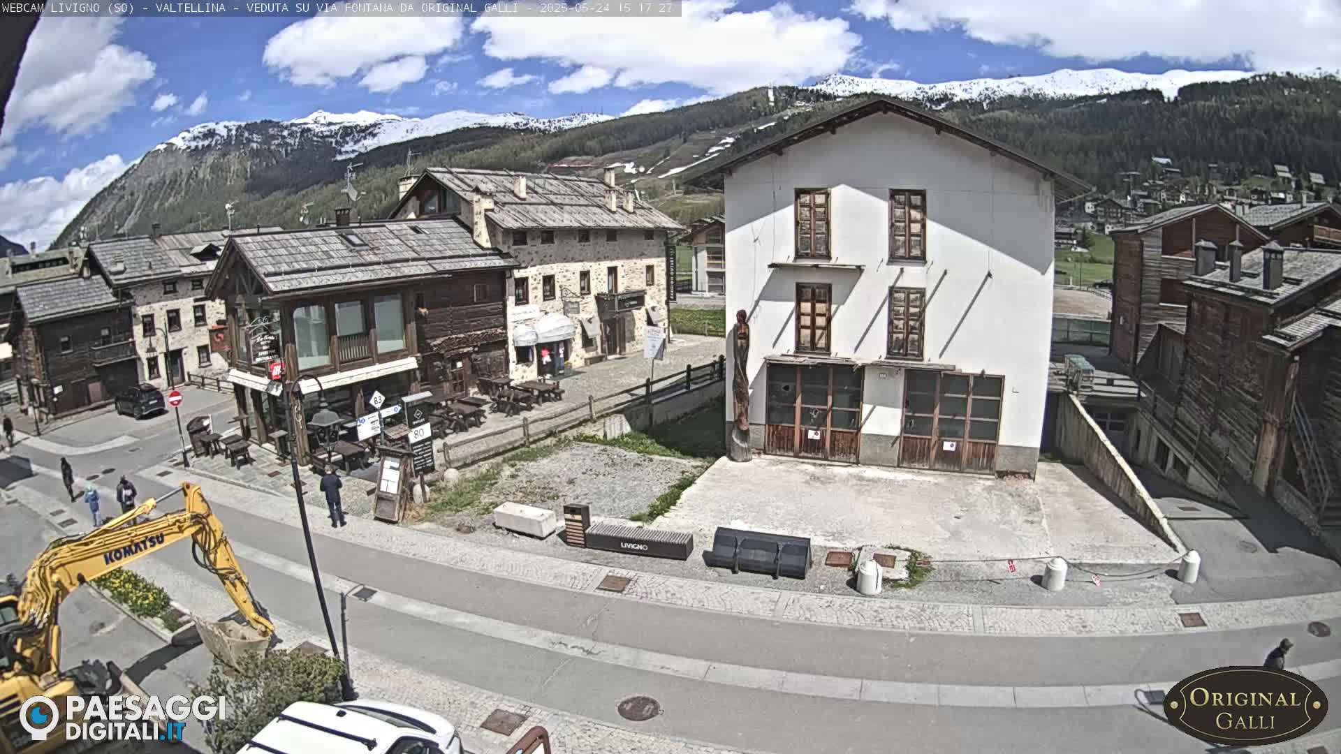 A sunny day in a mountain village, showing several buildings, a road, a construction vehicle, and snow-capped mountains in the background.
