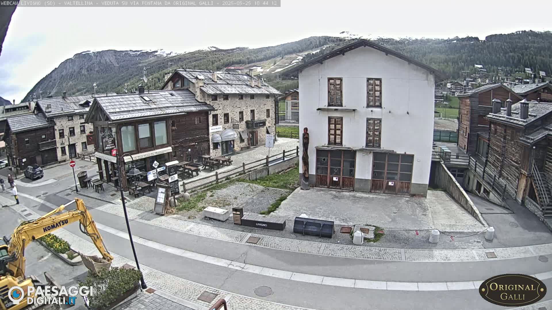 A mostly sunny view of a town square in a mountain village, with a yellow excavator parked on the street and several buildings visible, including some made of stone and wood.