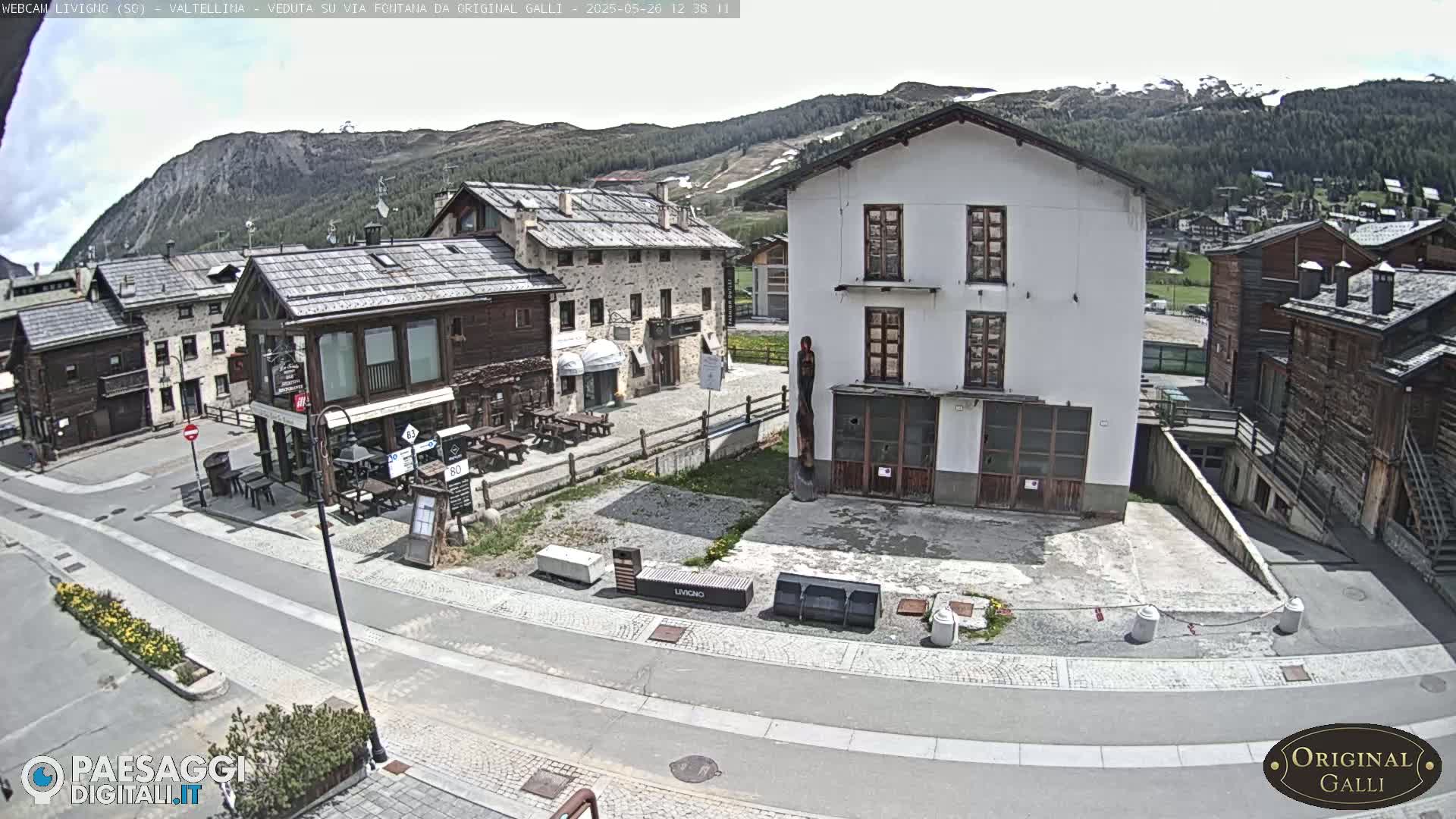 A mostly sunny view of a village square with several buildings, a paved road, and some outdoor seating.