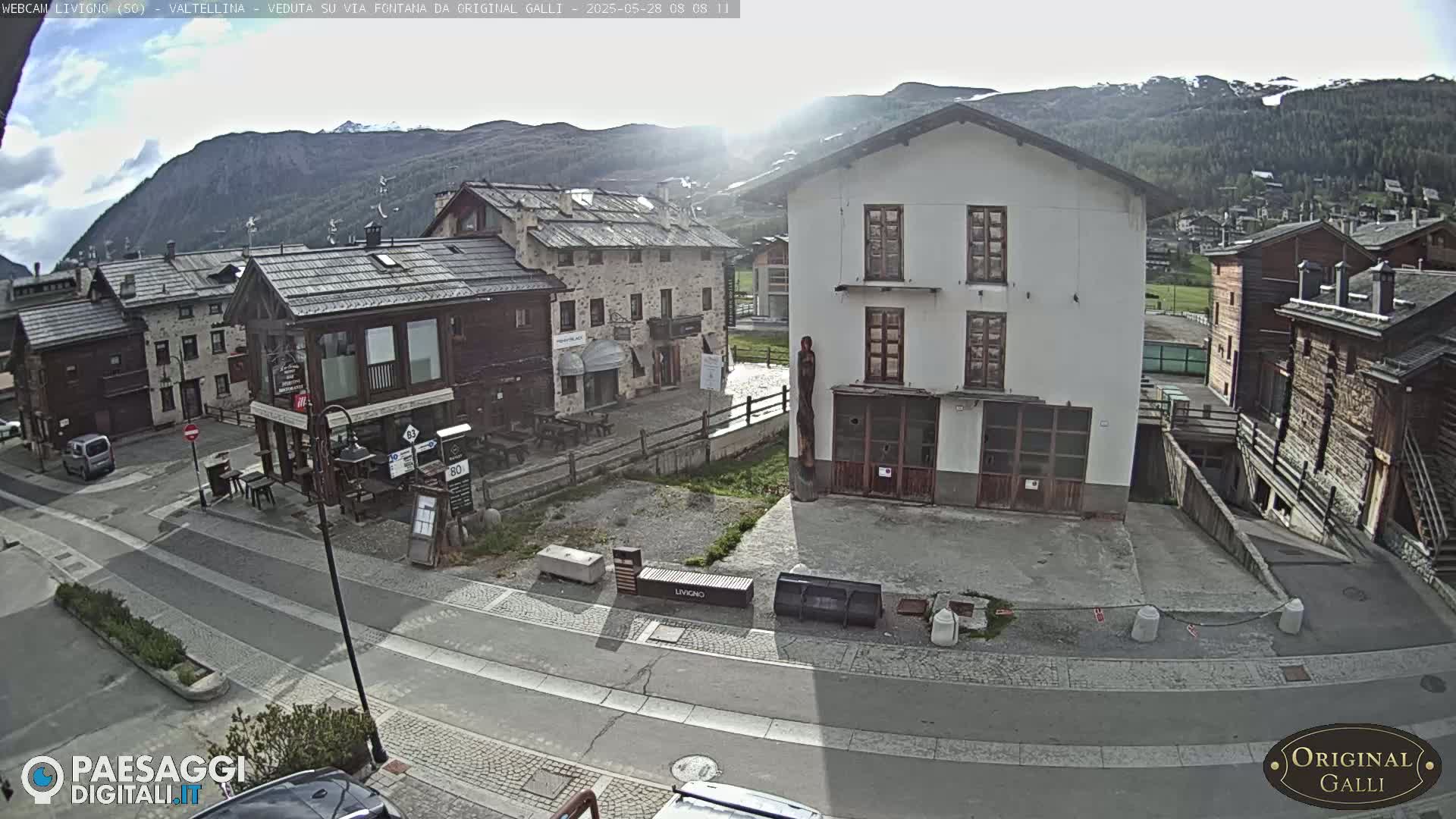 A sunny day in a village nestled in a valley surrounded by mountains shows a street scene with several buildings, including a stone building with wooden accents and a tall white building.