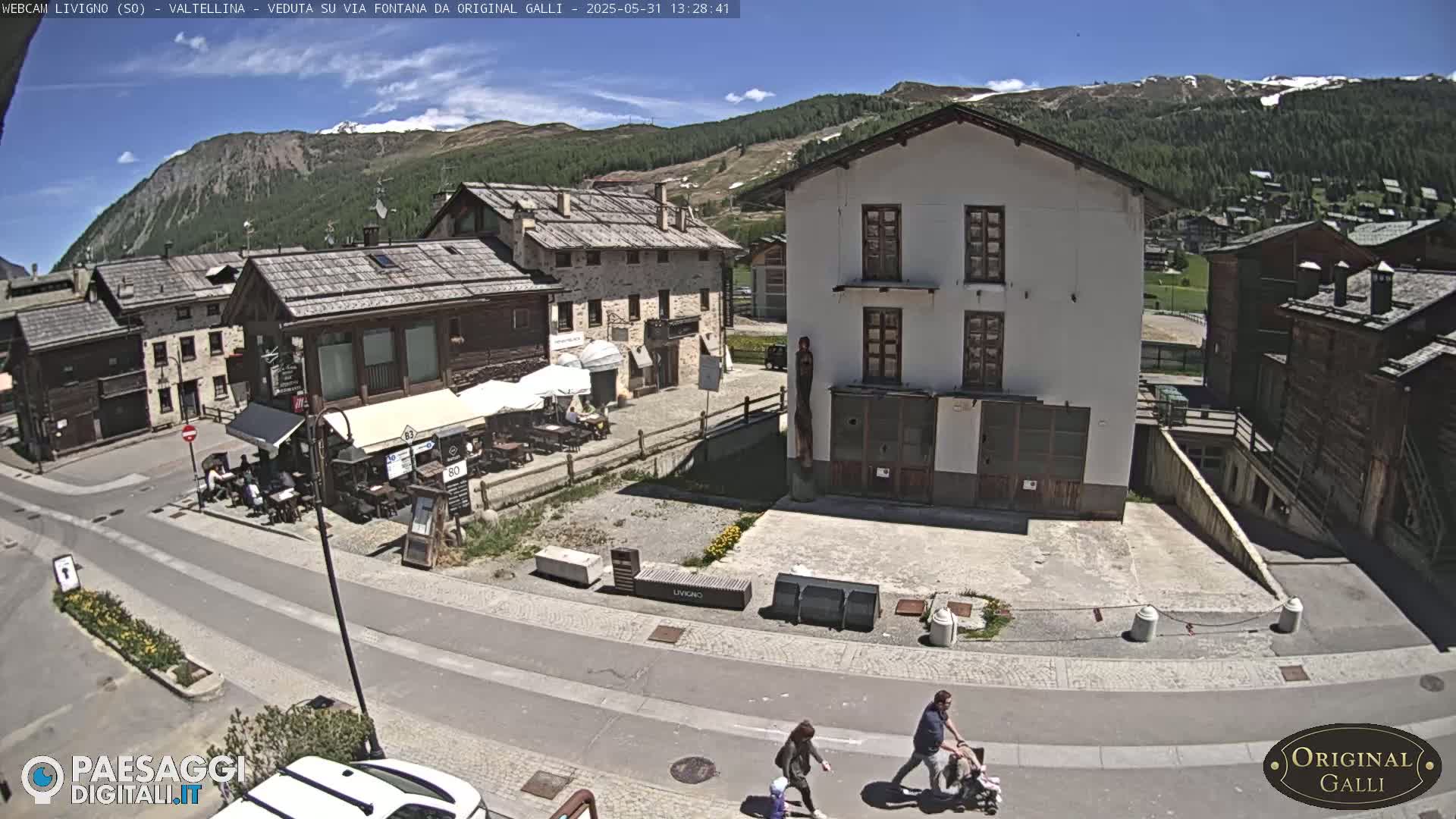 A sunny mountain village square with several buildings, a cafe with outdoor seating, and a few pedestrians.