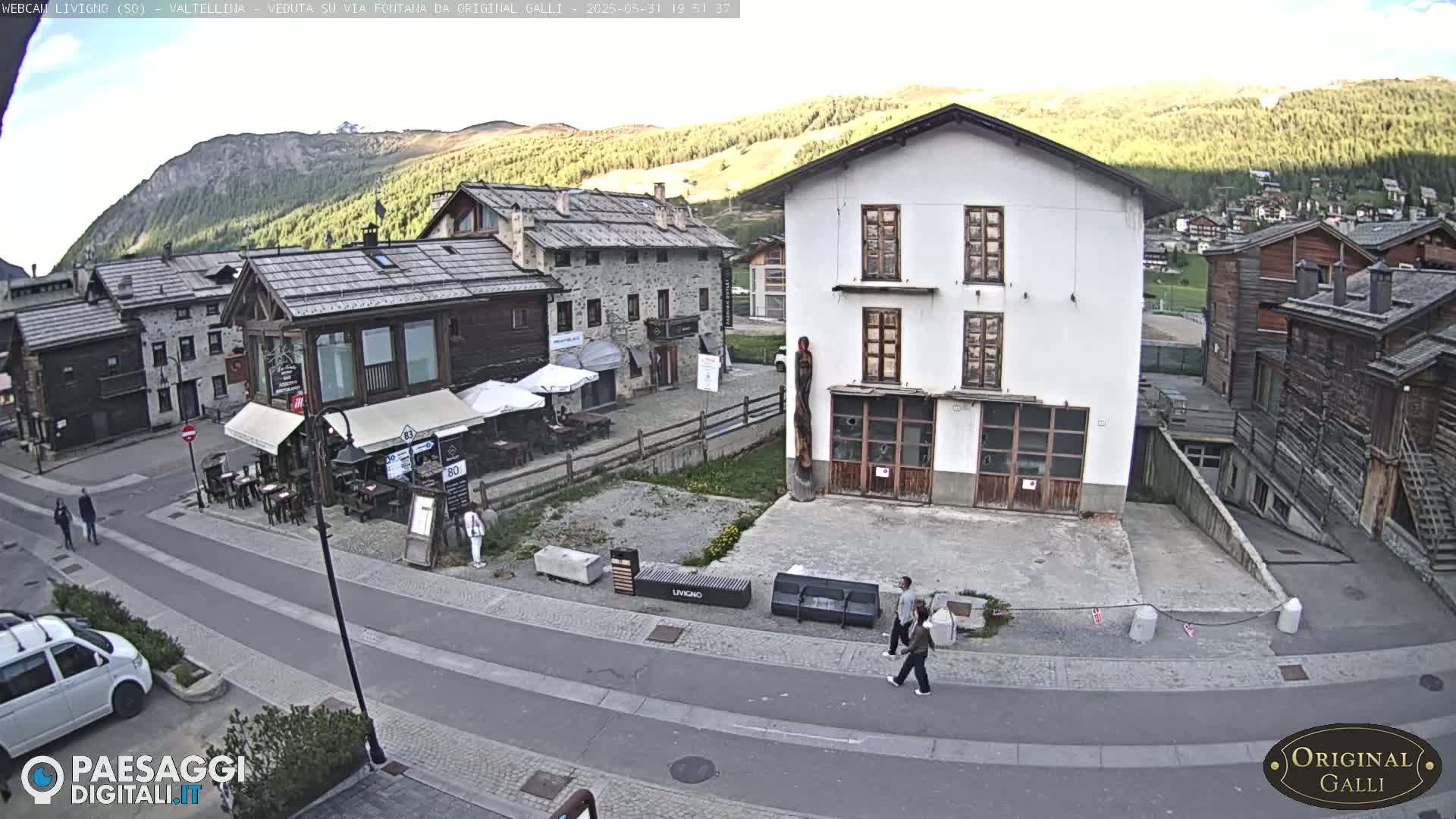 A mostly sunny view of a village square in the mountains, with several buildings, a small outdoor cafe, and a few pedestrians.