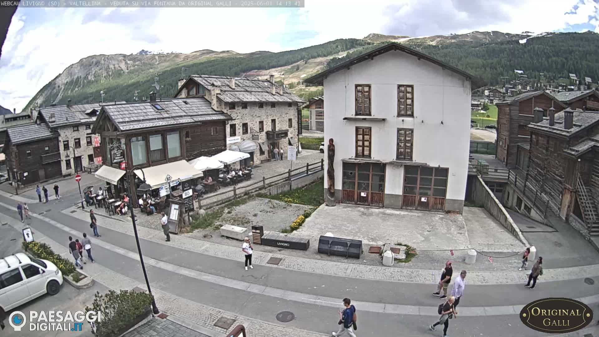 A partly cloudy day shows a village street scene with pedestrians, cars, and buildings in a mountainous region.