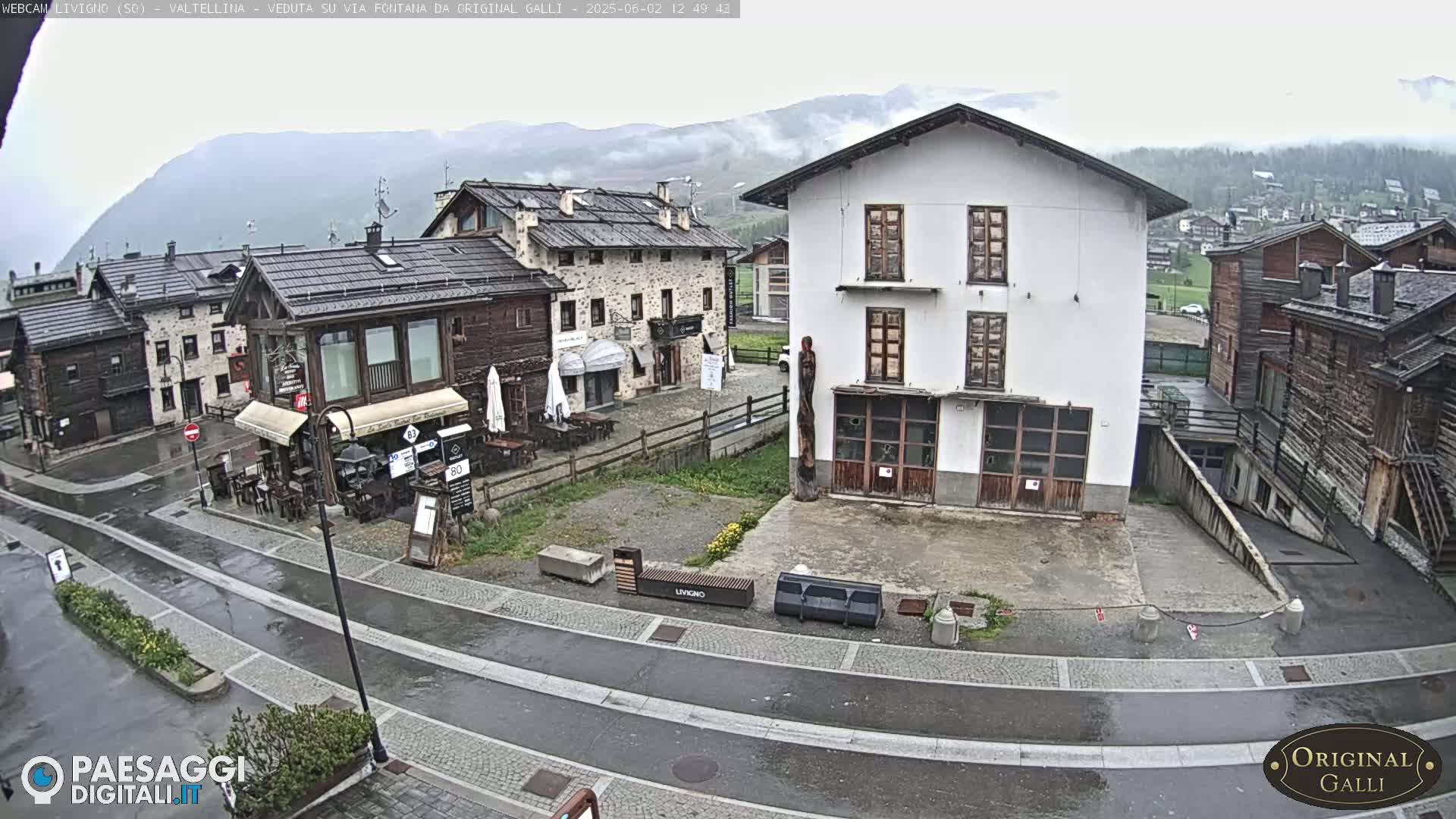 A wet, village square in the mountains features several stone and wood buildings, a few benches, and a mostly empty road.