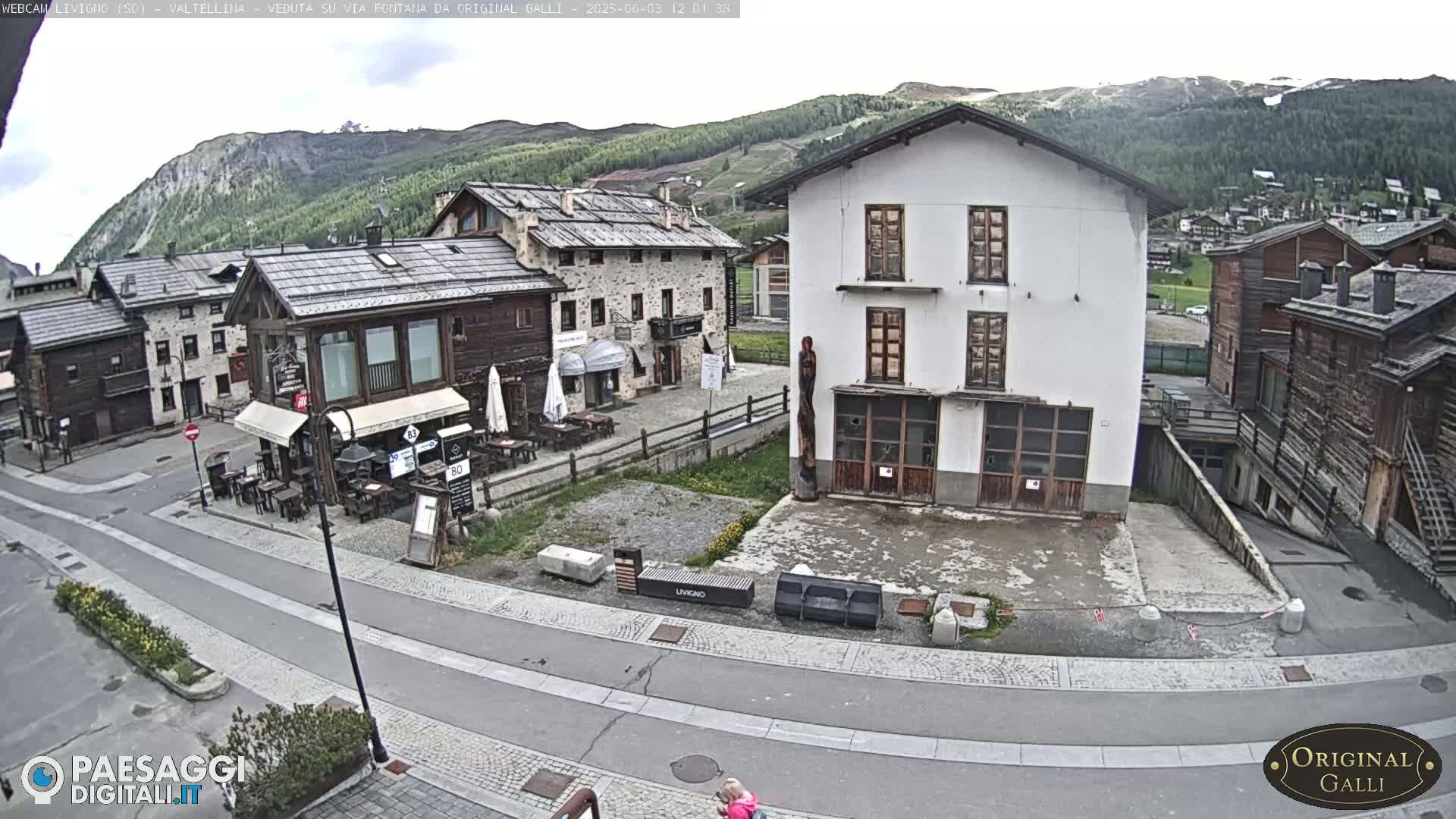 A mostly cloudy day in a small European village with stone and wood buildings, a paved road, and some outdoor seating in front of a cafe.