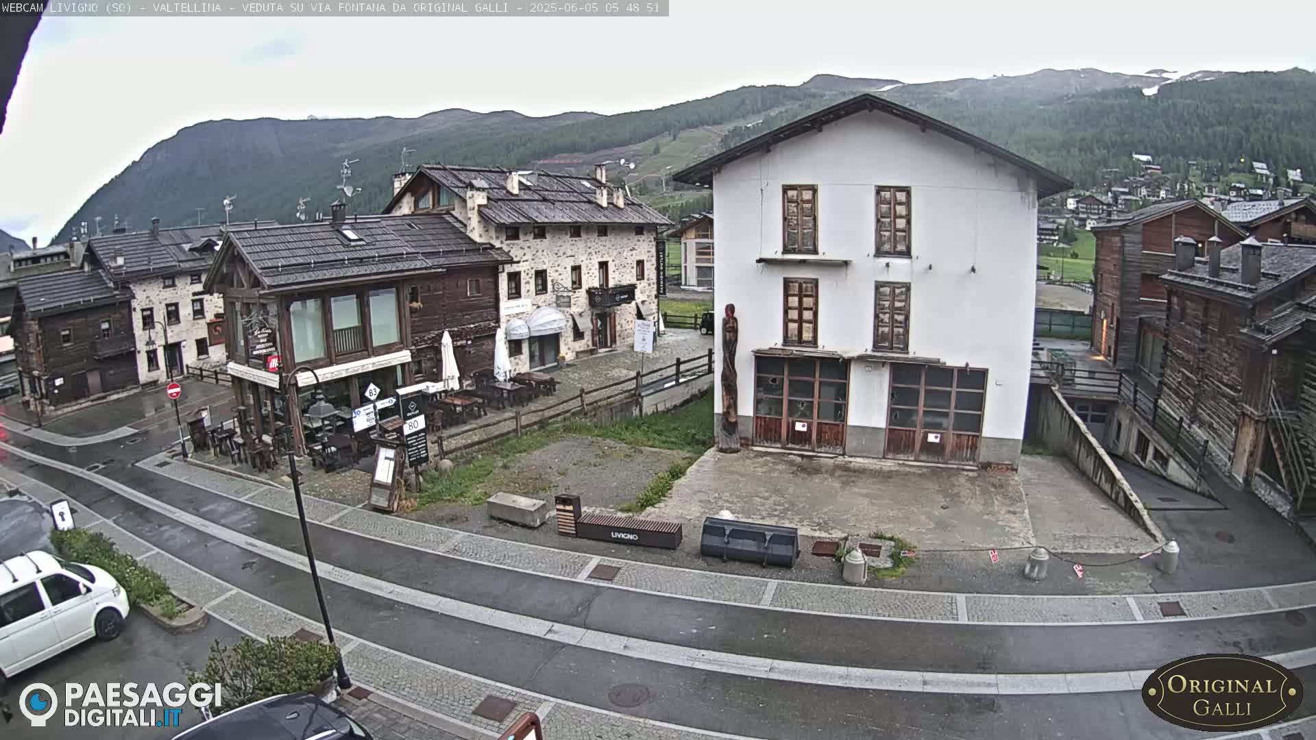 A wet village square with several buildings, including a restaurant and a larger white building, is nestled in a valley with mountains in the background under an overcast sky.