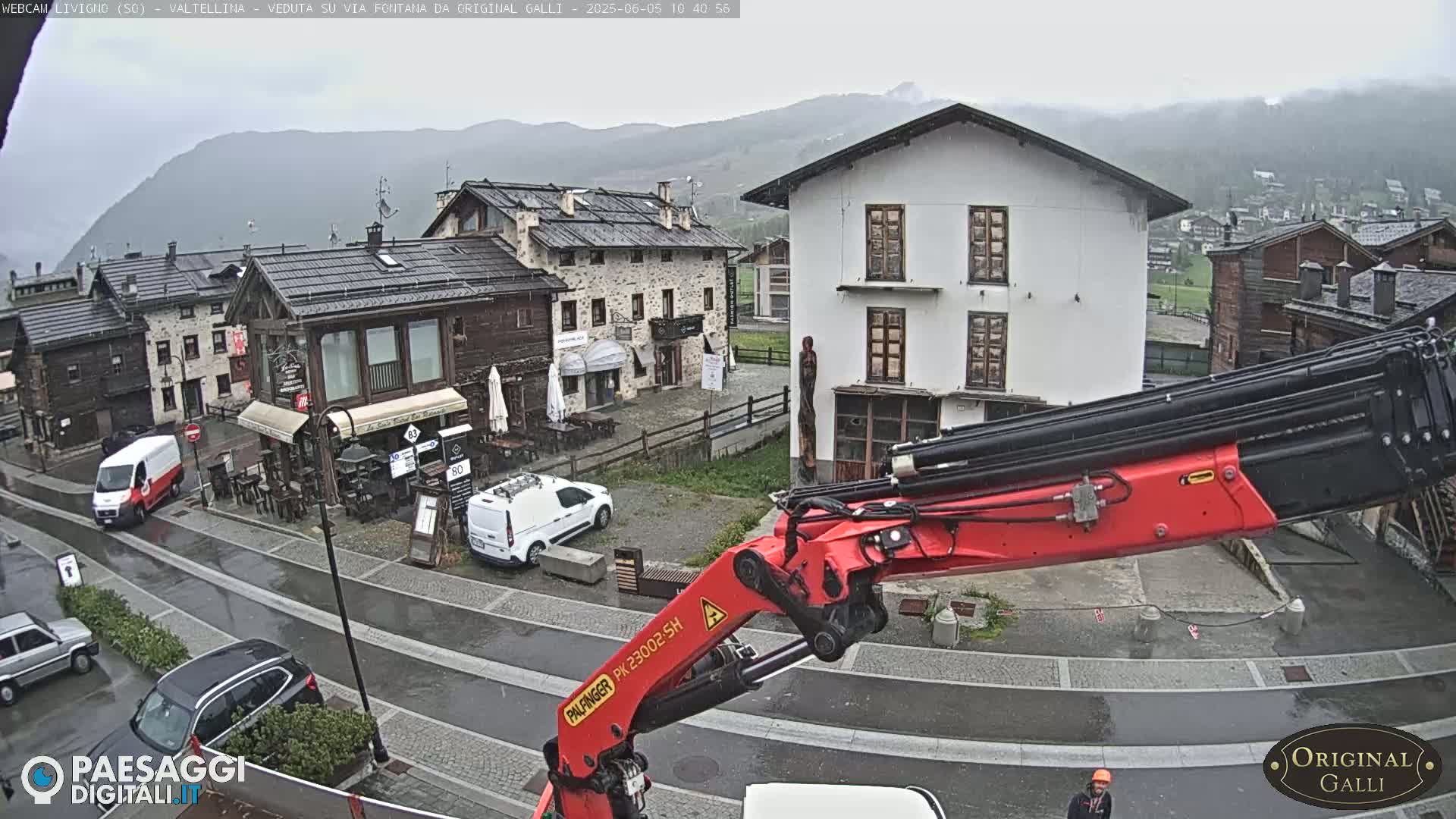 A red crane arm dominates the foreground of a rainy village scene nestled in a valley, with several cars parked along wet streets and stone buildings in view.