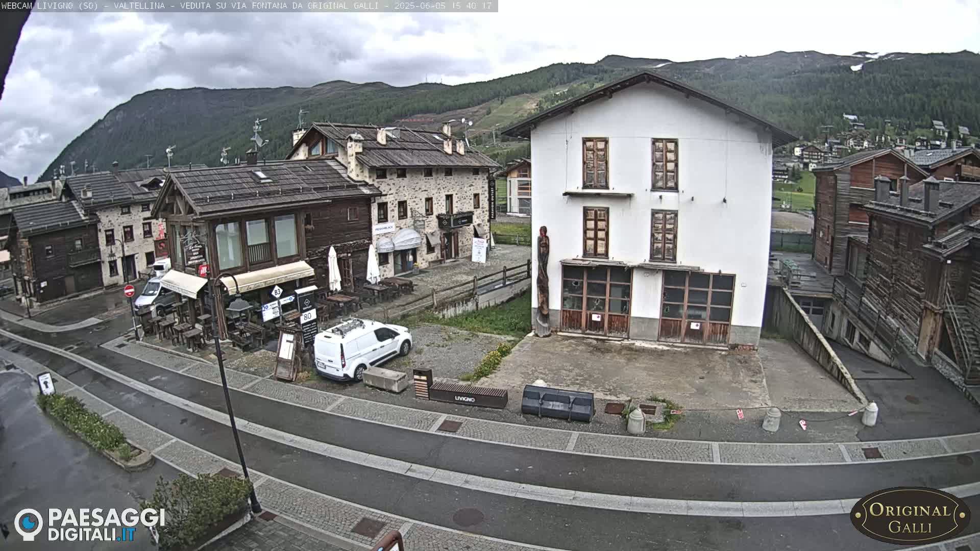 A wet, overcast day in a small village nestled in a mountain valley, showing several buildings and a sparsely populated street.