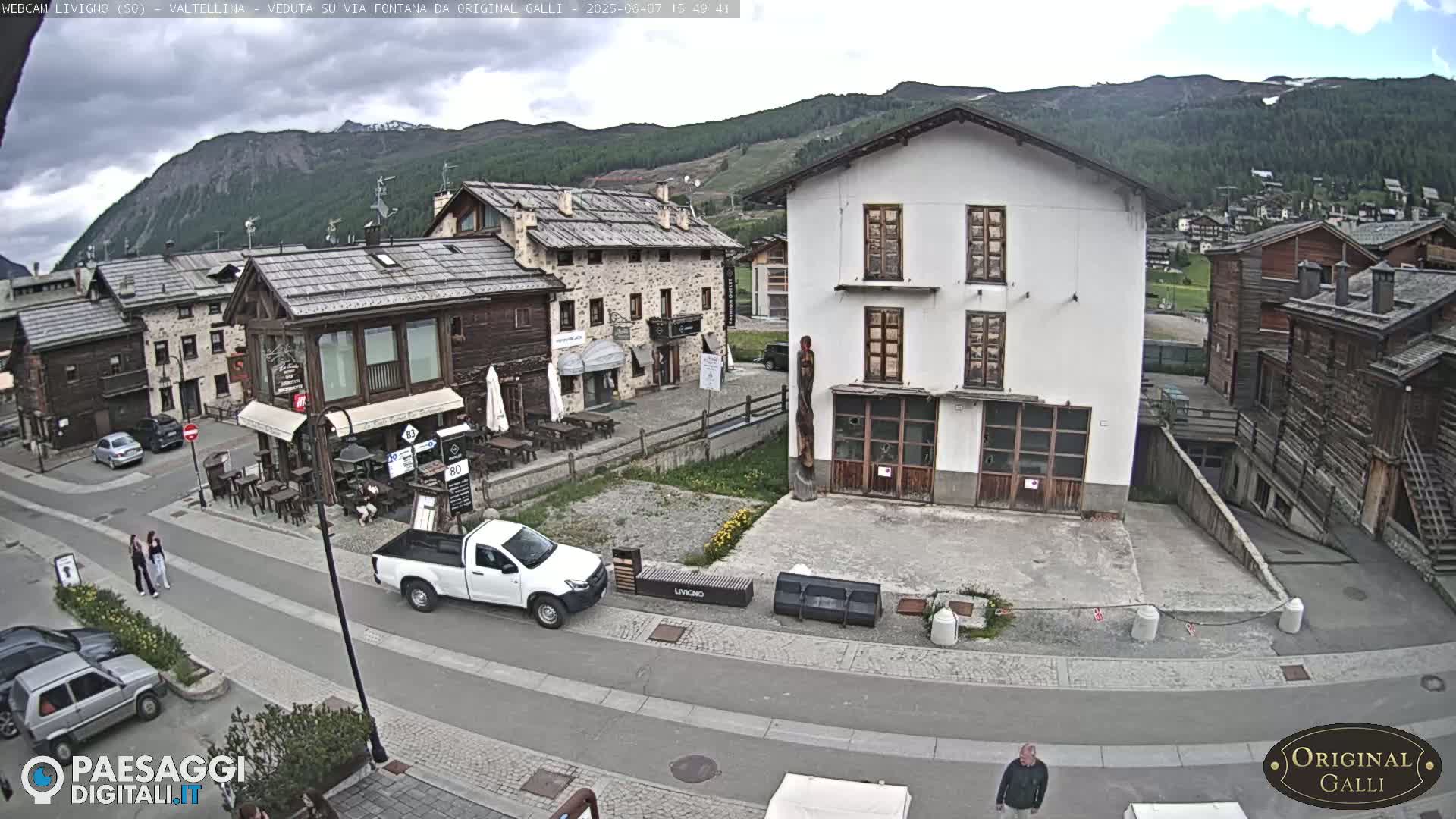 A partly cloudy day in a mountainous village with several parked cars, a white pickup truck, and a few people walking, situated around buildings of varying architectural styles.