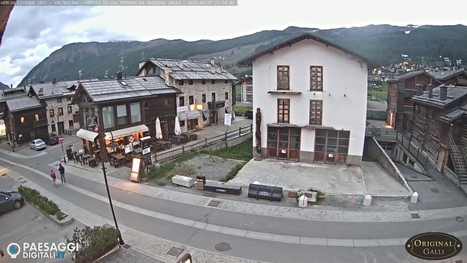 A mostly empty village square in the mountains under an overcast sky, with several buildings and a few people walking.