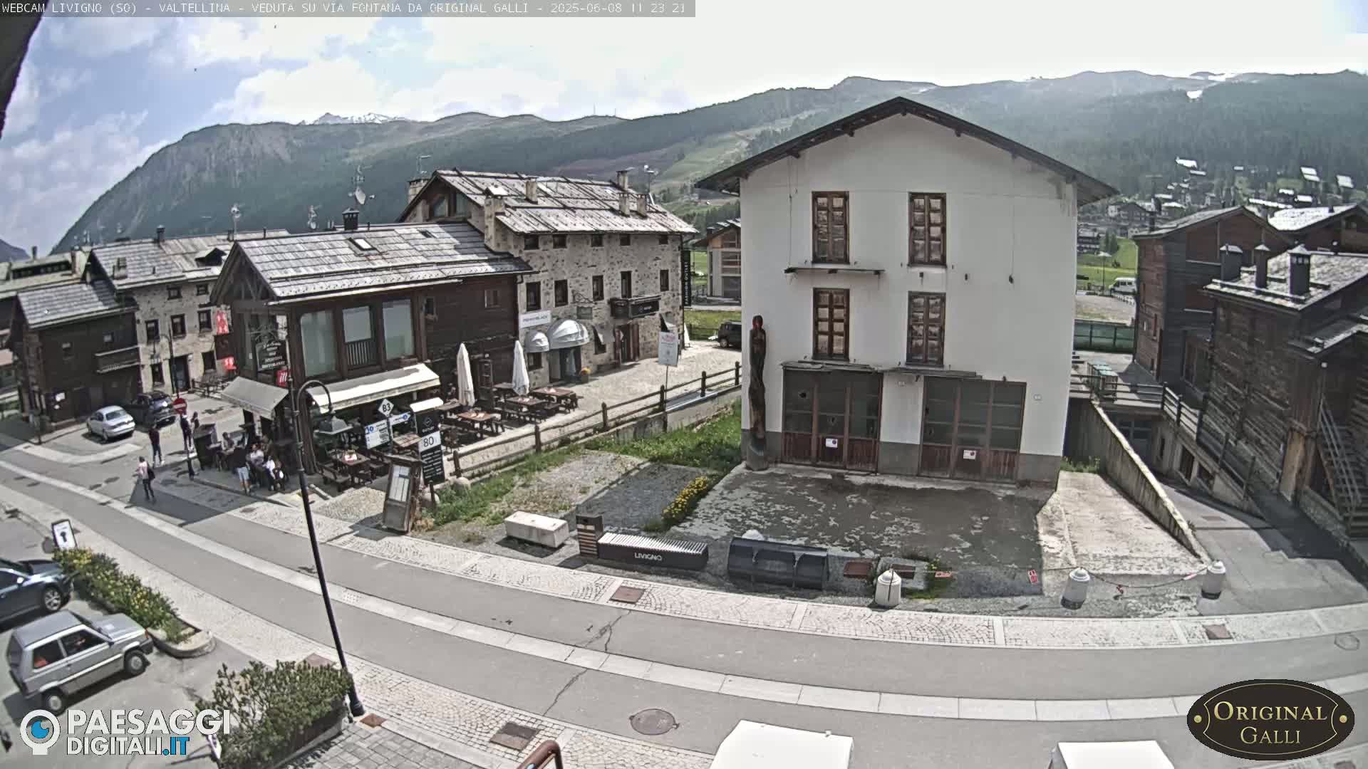 A sunny day in a village nestled in a valley surrounded by mountains, showing a street with parked cars and buildings, including a restaurant with outdoor seating.