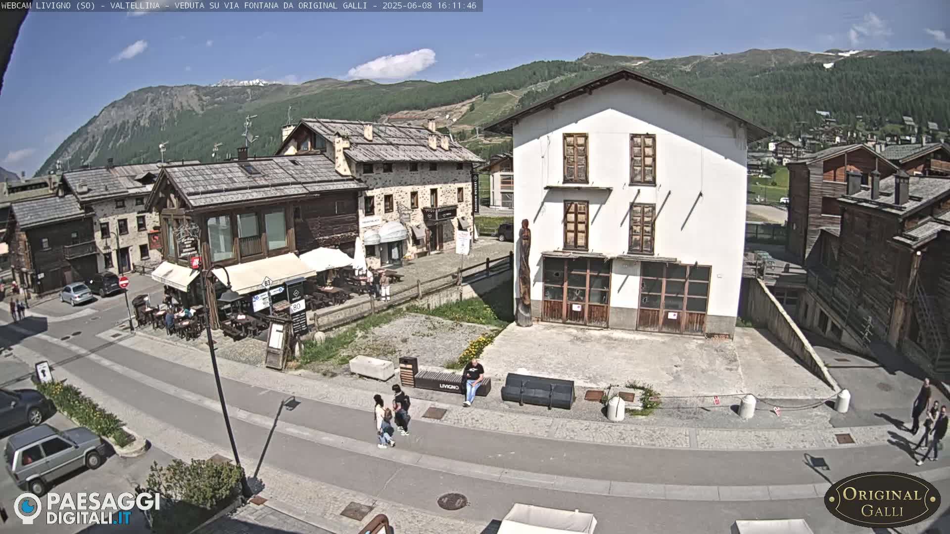 A sunny day in a village nestled in a valley amongst mountains, showing a street with cars, buildings, and a few people.
