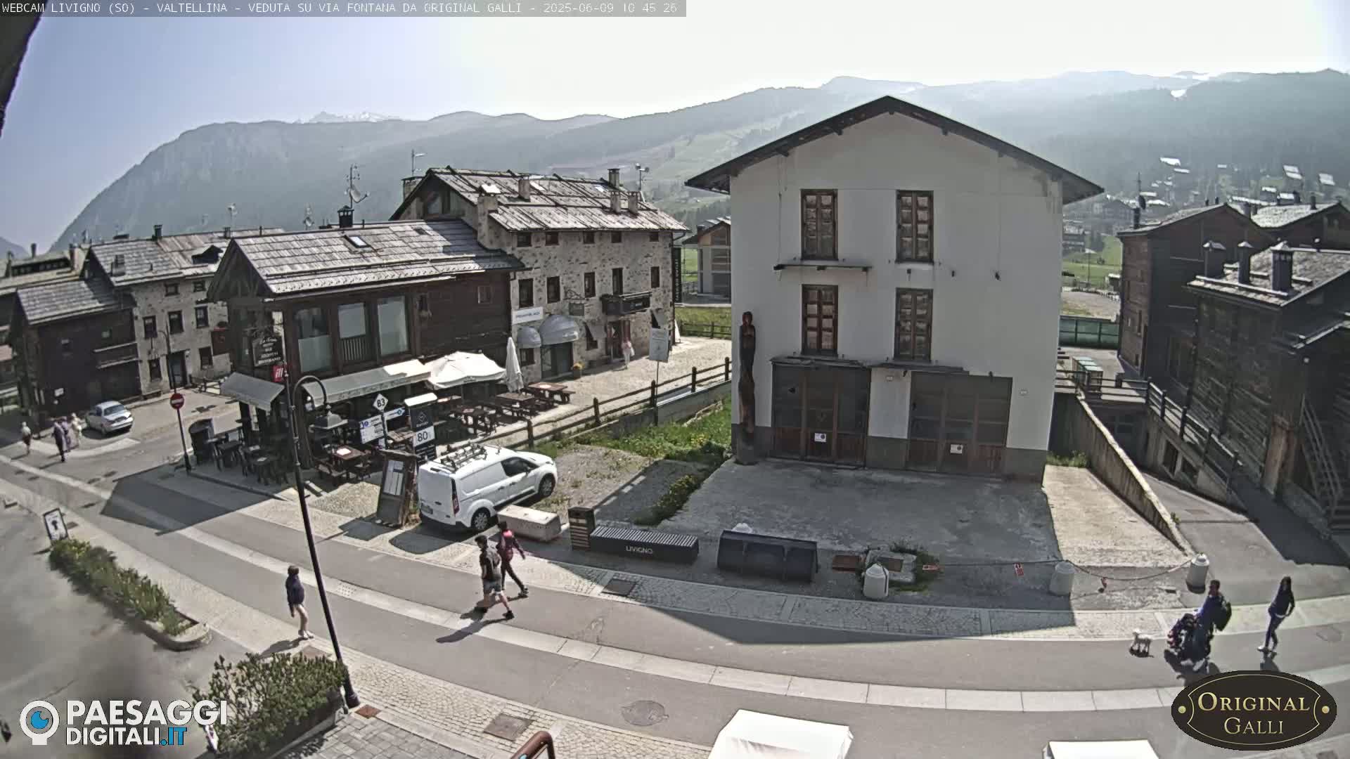 A sunny day in a mountainous village shows a paved square with several buildings, a few people walking, and a few parked cars.
