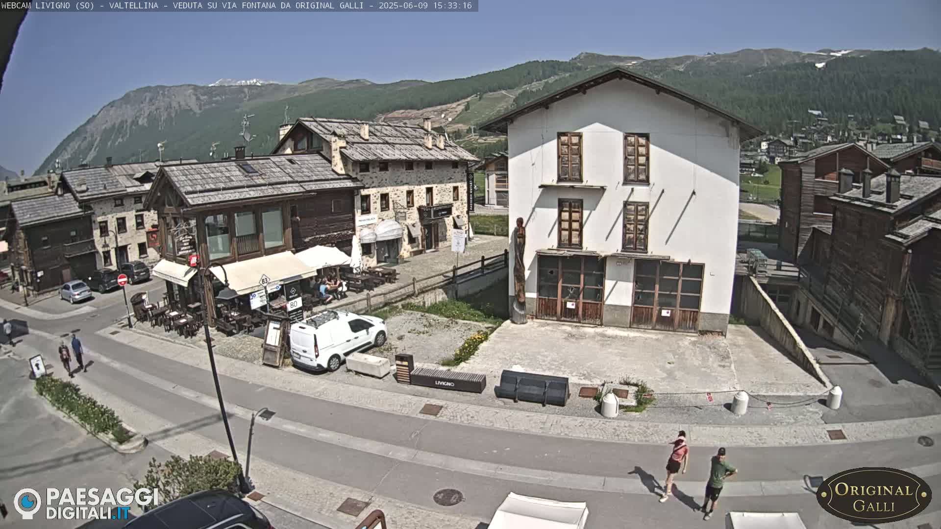 A sunny mountain village square is viewed from above, showing several buildings, parked cars, a few pedestrians, and outdoor seating at a restaurant.