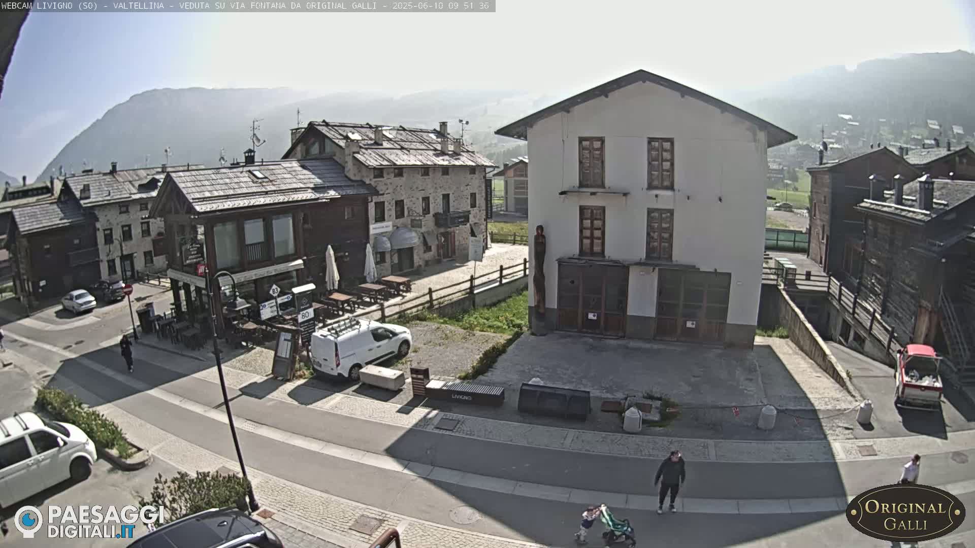 A sunny view of a village street scene with several buildings, a few cars, and pedestrians, situated in front of a mountain range.