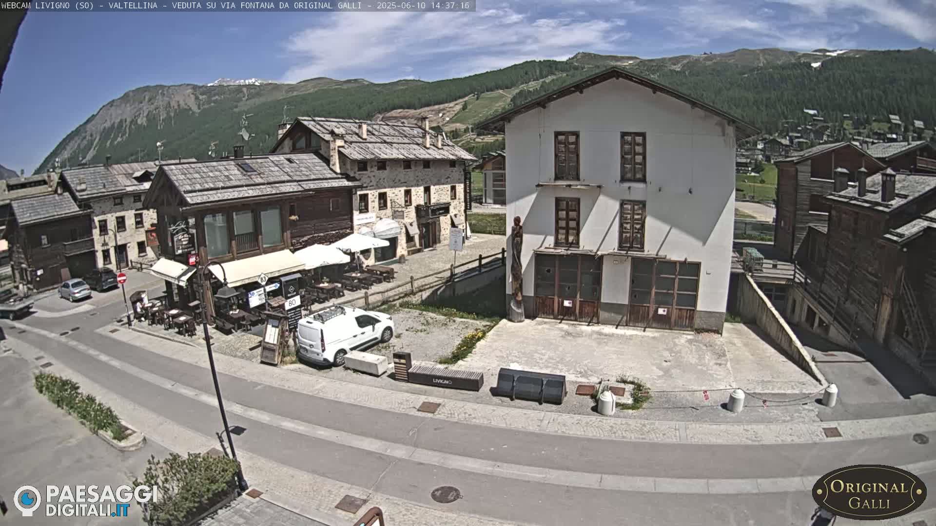 A sunny view of a village street with several buildings, a few parked cars, and outdoor seating at a restaurant, nestled in a mountain valley.